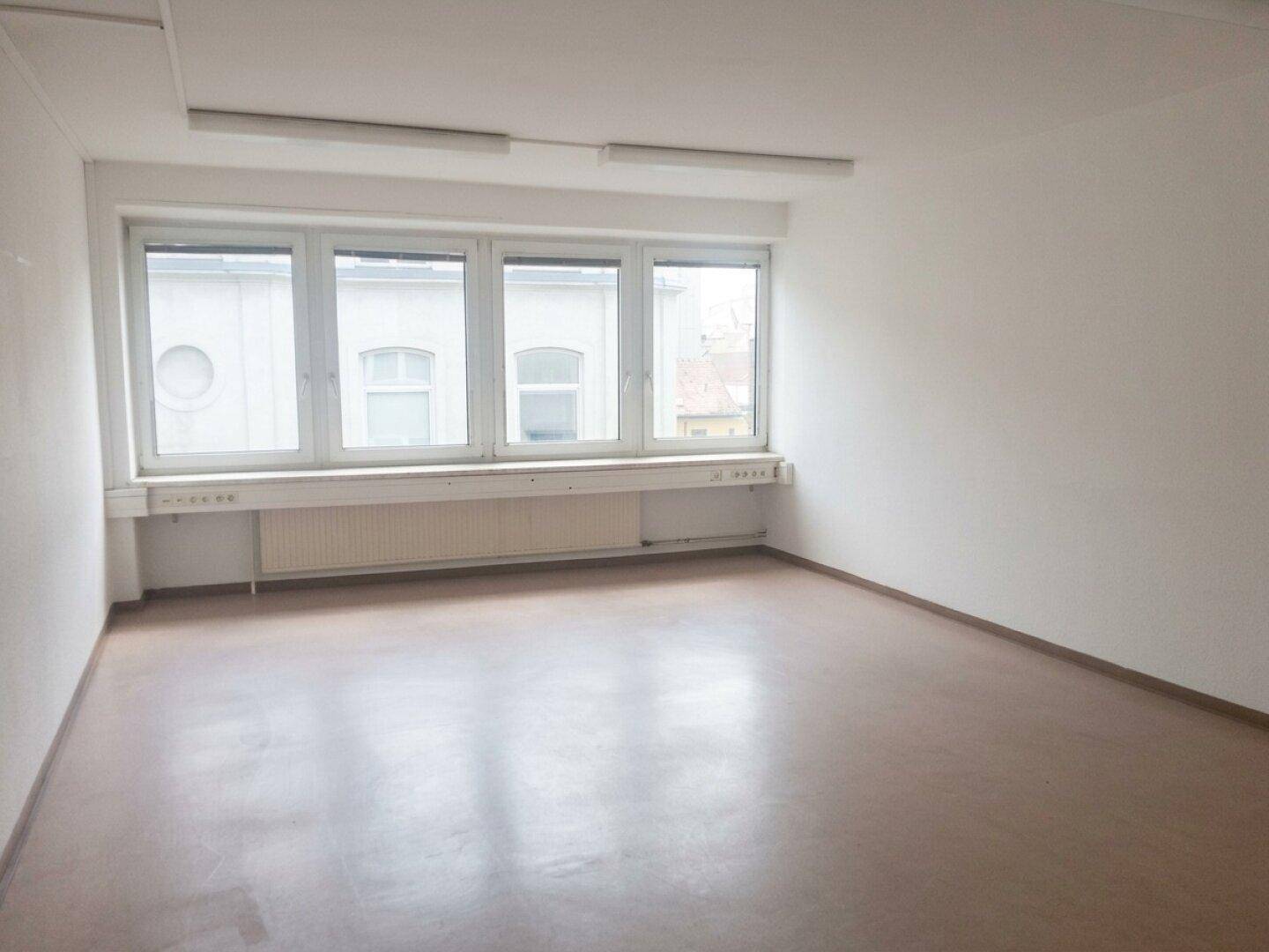 Heller, leerer Raum mit großen Fenstern und Heizkörper unter der Fensterbank, ideal für ein Büro.