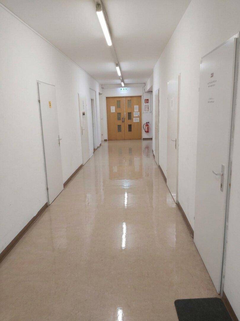 Langer, heller Flur mit vielen Türen und glänzendem Bodenbelag, führt zu einem Ausgang mit Doppeltür.