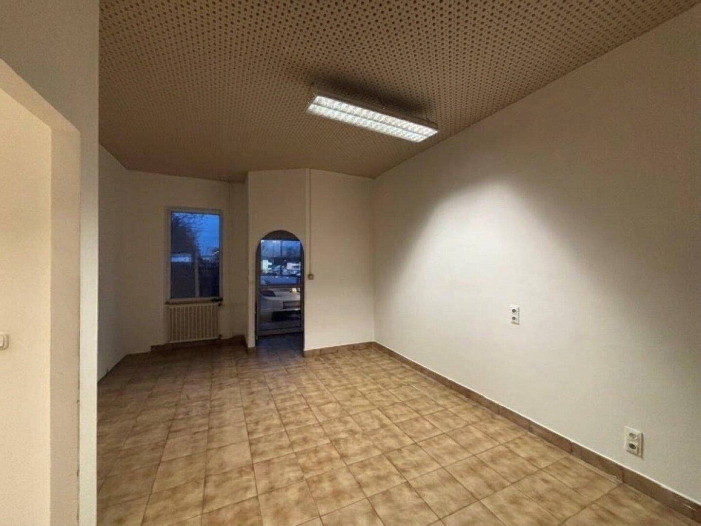 Heller Büroraum mit braunen Fliesen, Fenster und abgehängter Decke mit Akustikplatten.