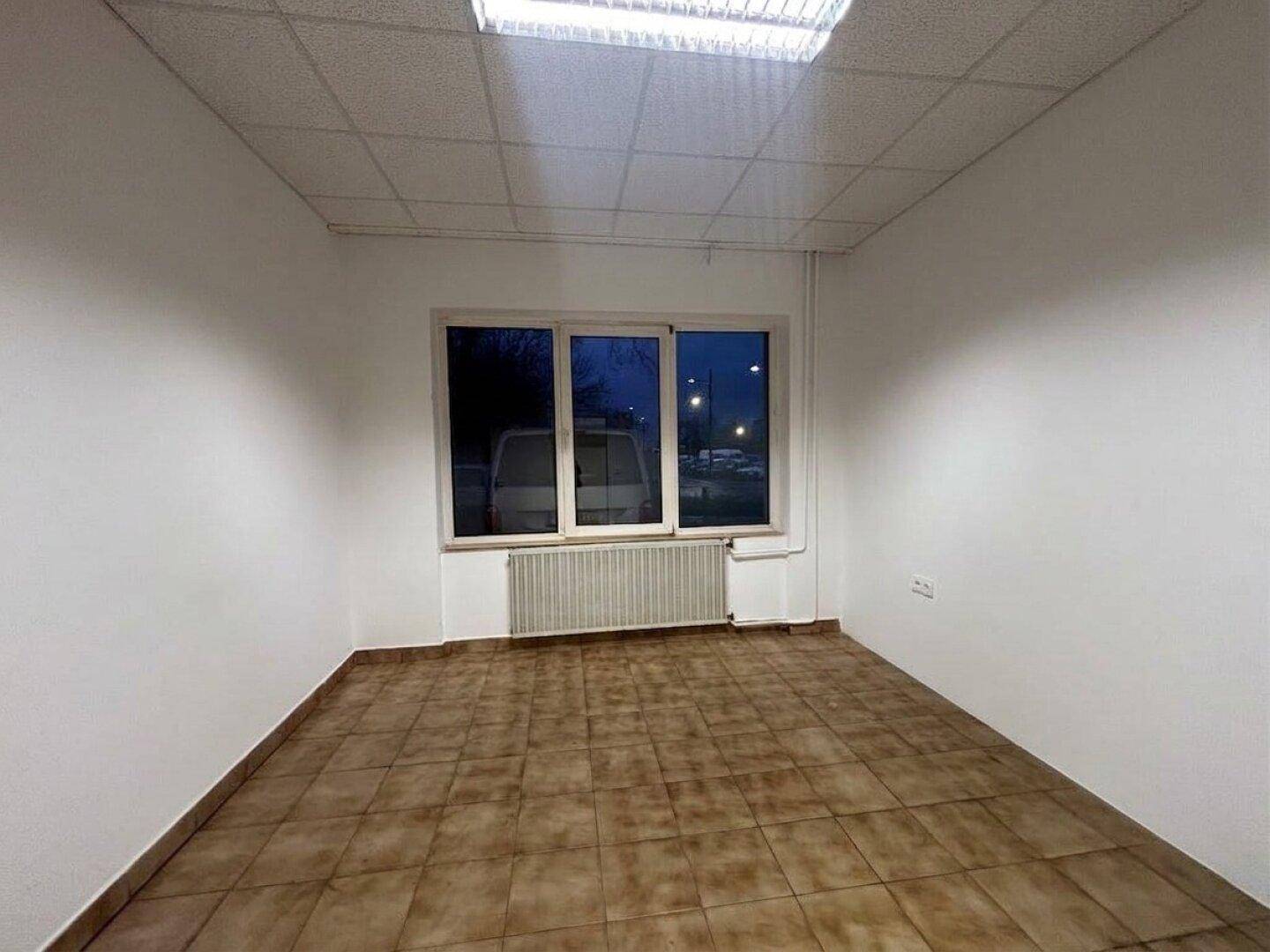 Büroraum mit großen Fenstern, braunen Fliesen und abgehängter Decke, gut beleuchtet.