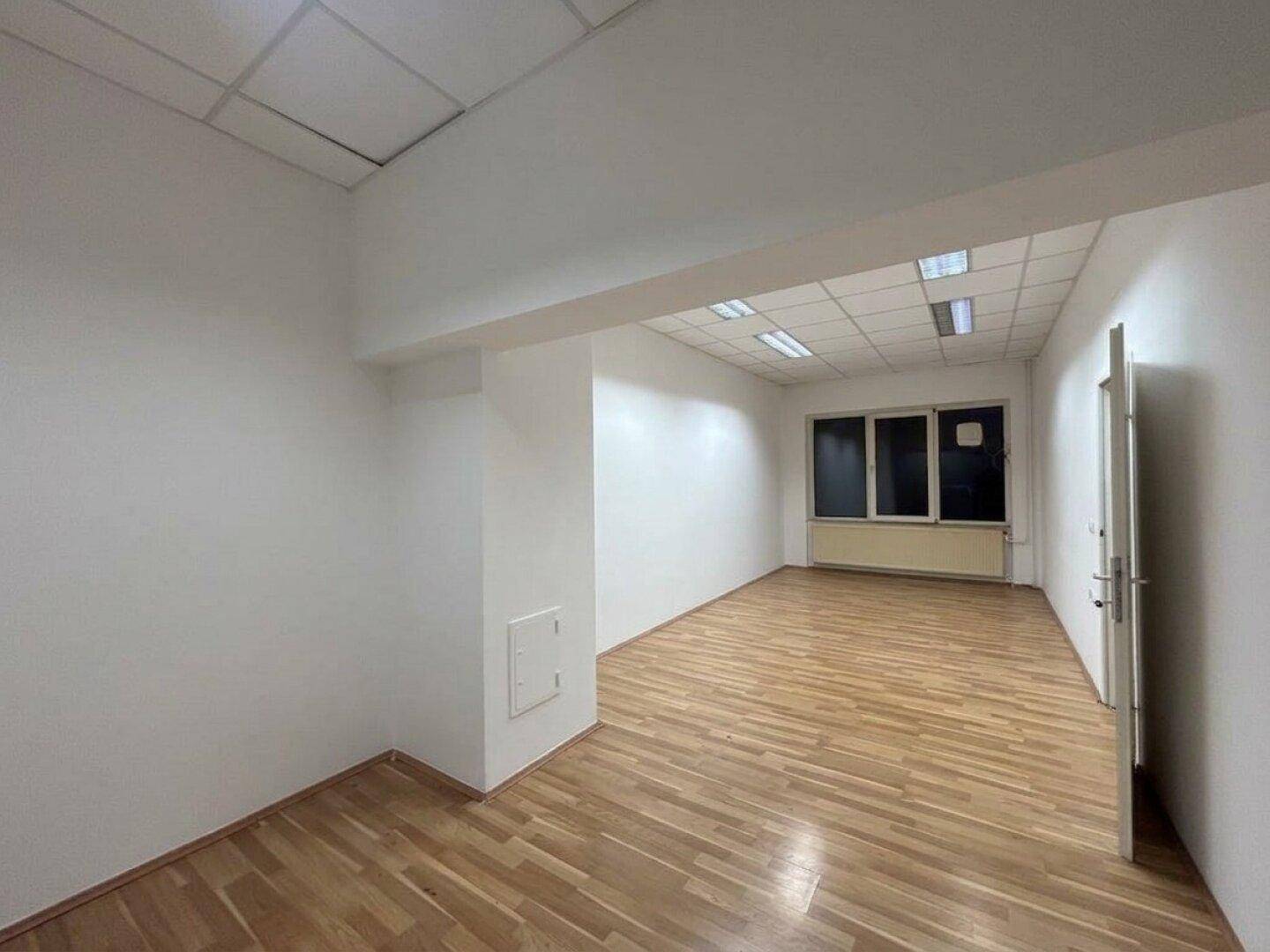 Langer Büroraum mit hellem Laminatboden, Fenster und abgehängter Decke.