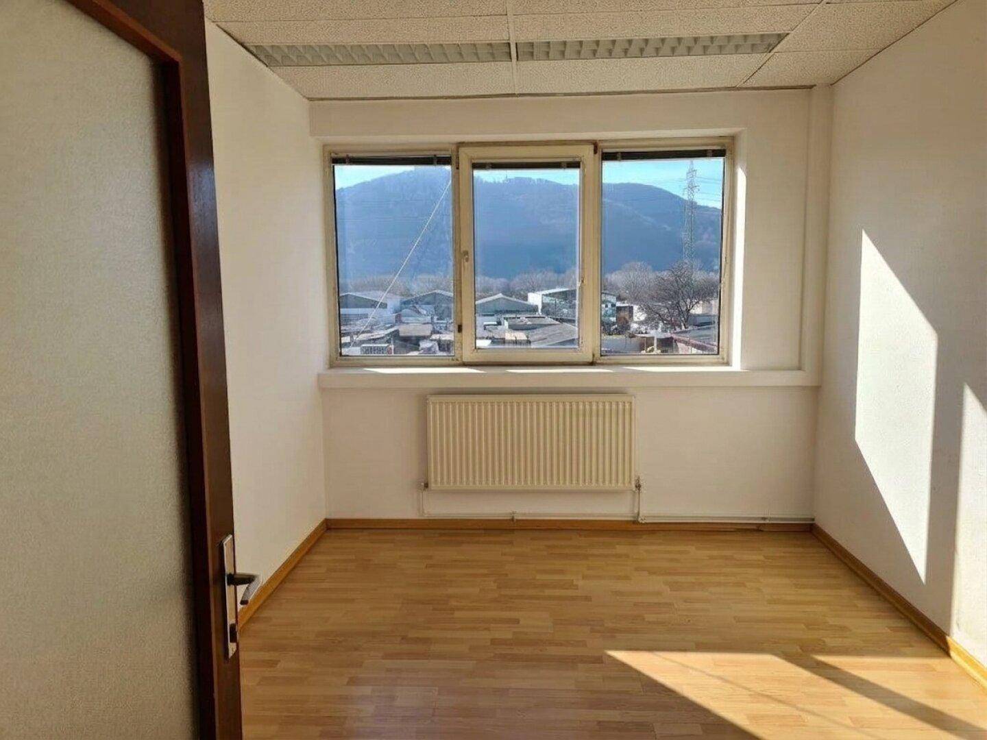 Helles, leeres Büro mit Holzboden, Heizkörper und Fenster mit Bergblick und viel Tageslicht.