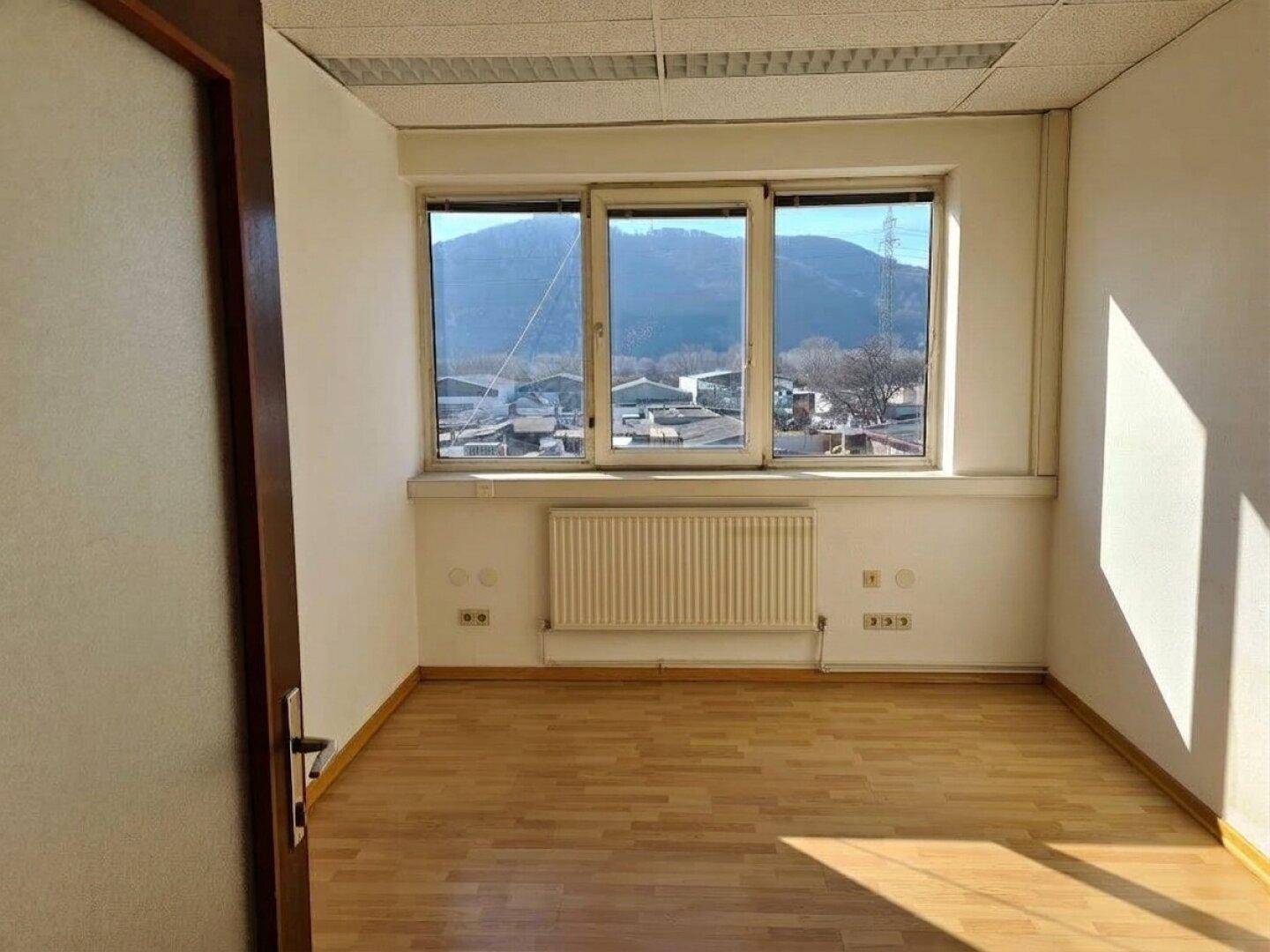 Helles, leeres Zimmer mit Laminatboden, weißen Wänden und einem großen Fenster mit Bergblick.