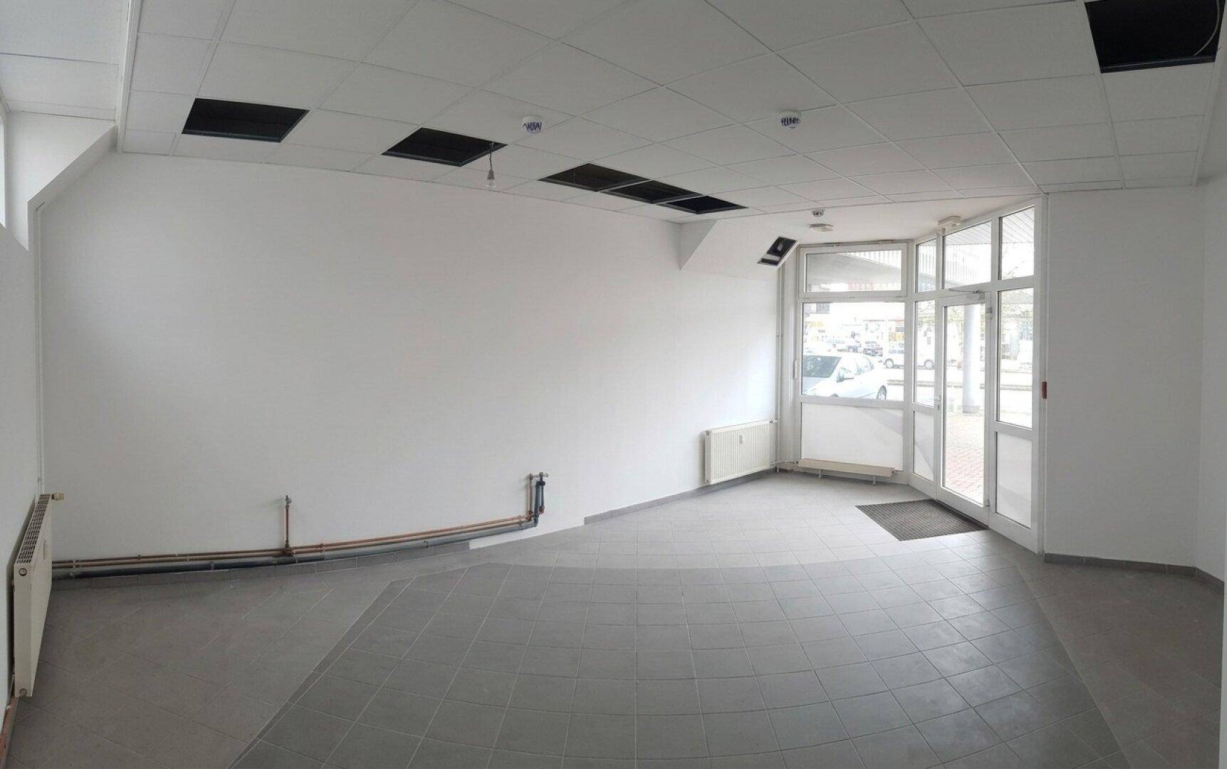 Großer, leerer Gewerberaum mit Fliesenboden, Heizkörpern und einer Glasfront zum Eingangsbereich.