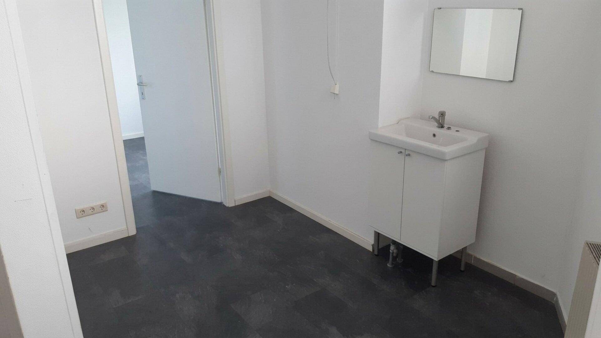 Kleiner Waschraum mit Waschbecken, Spiegel und dunklem Bodenbelag, ausgestattet mit Heizkörper.