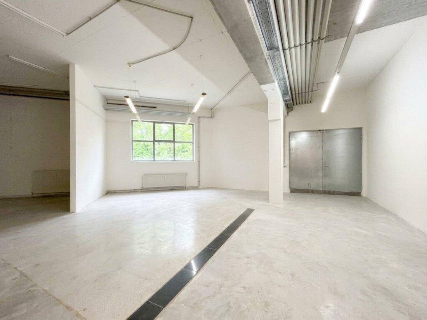 Heller Gewerberaum mit großen Fenstern, Betonboden und sichtbaren Lüftungsrohren an der Decke.