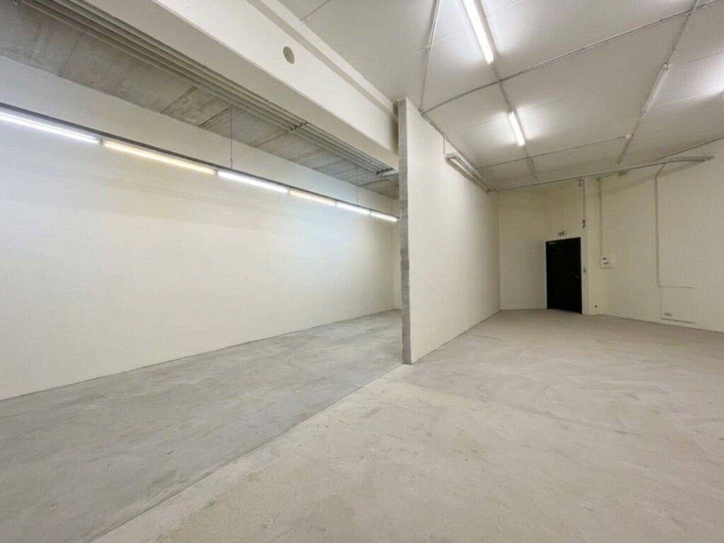 Geräumiger, leerer Gewerberaum mit Betonboden, hohen Decken und minimalistischer Beleuchtung.