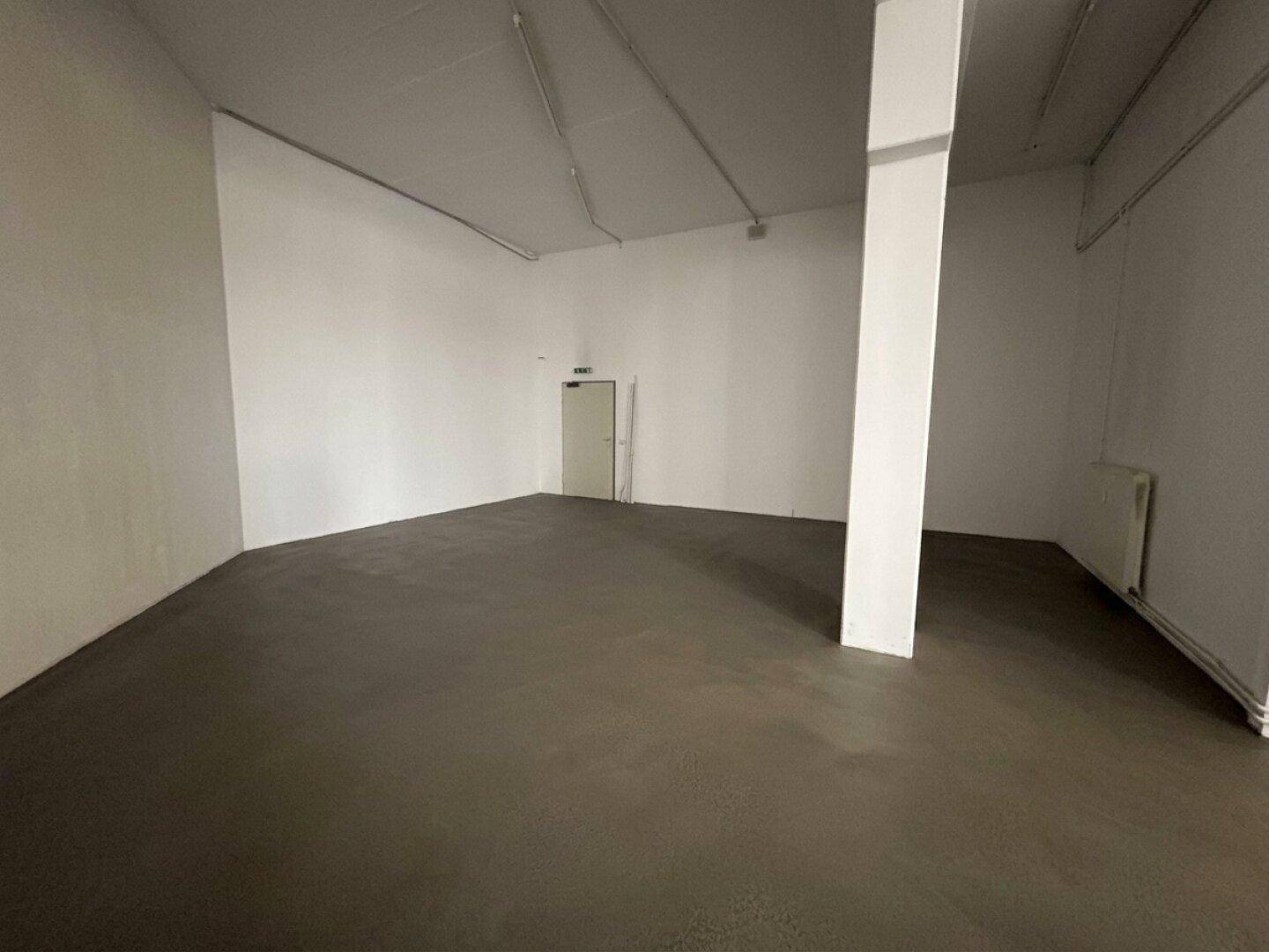 Geräumiger, leerer Gewerberaum mit dunklem Betonboden, weißen Wänden und sichtbaren Heizkörpern.