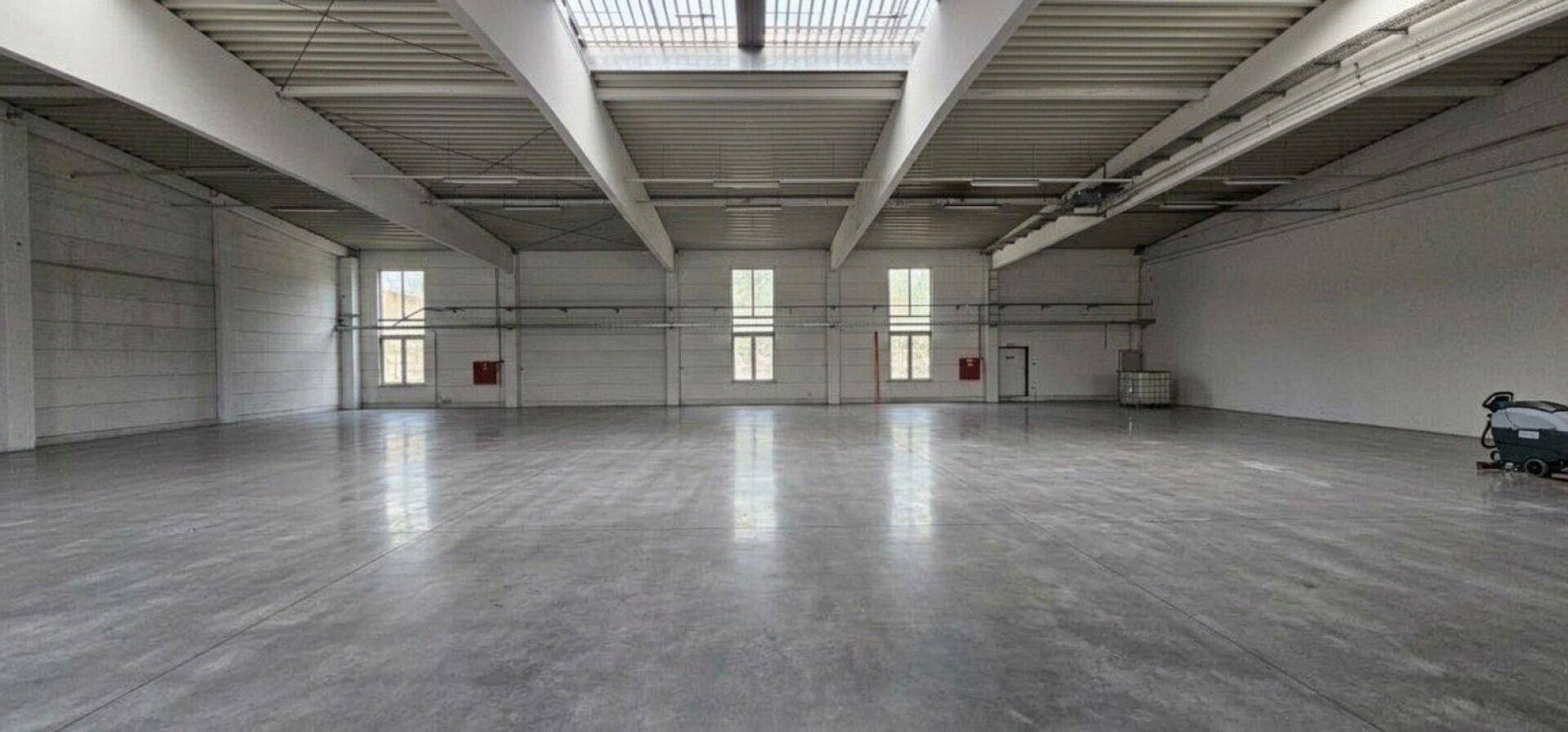 Große, helle Lagerhalle mit hohen Decken, Betonboden und Oberlichtern für viel Tageslicht.