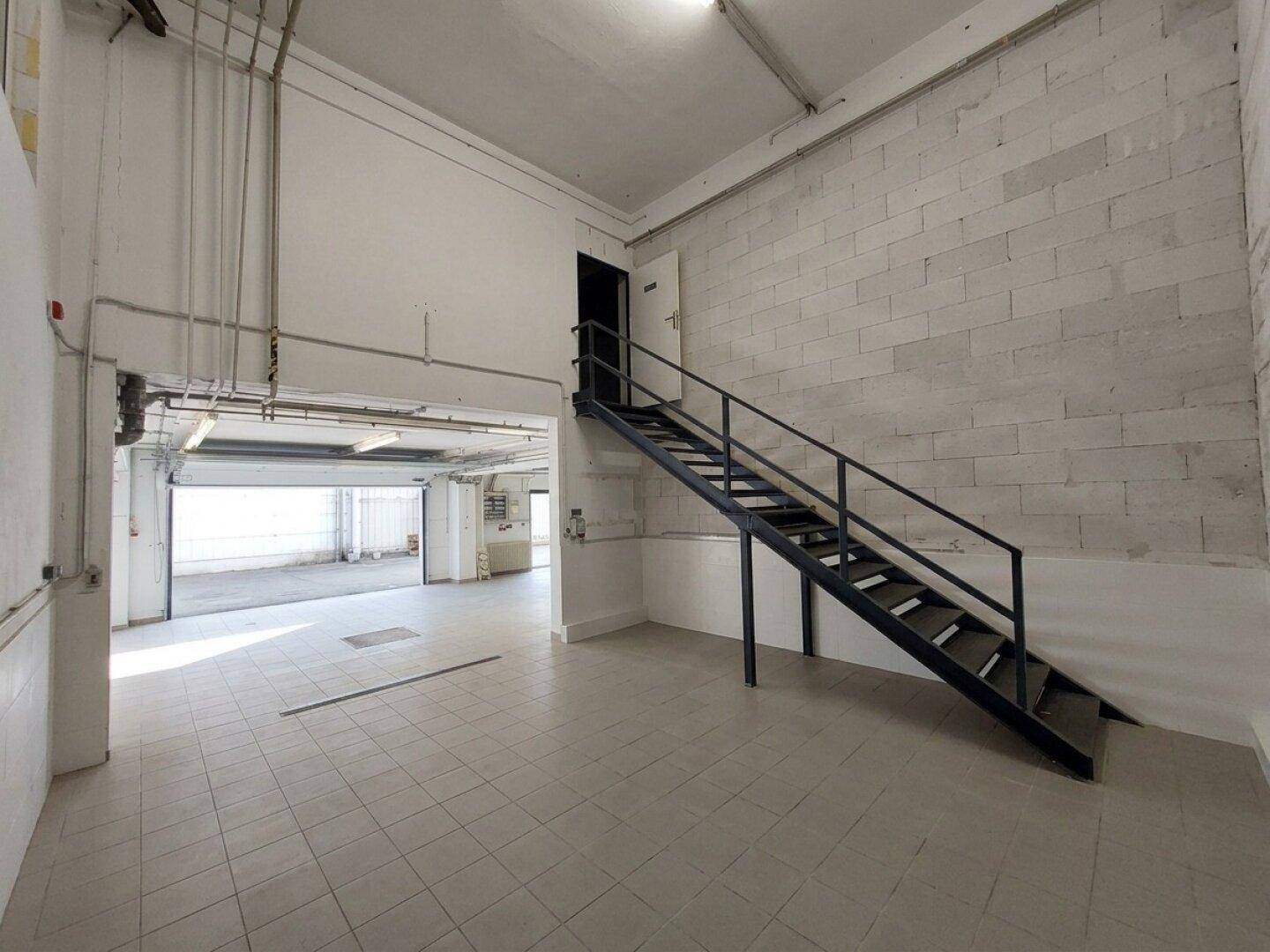 Großer, heller Gewerberaum mit Fliesenboden, einer Metalltreppe und offenem Zugang zu weiteren Bereichen.