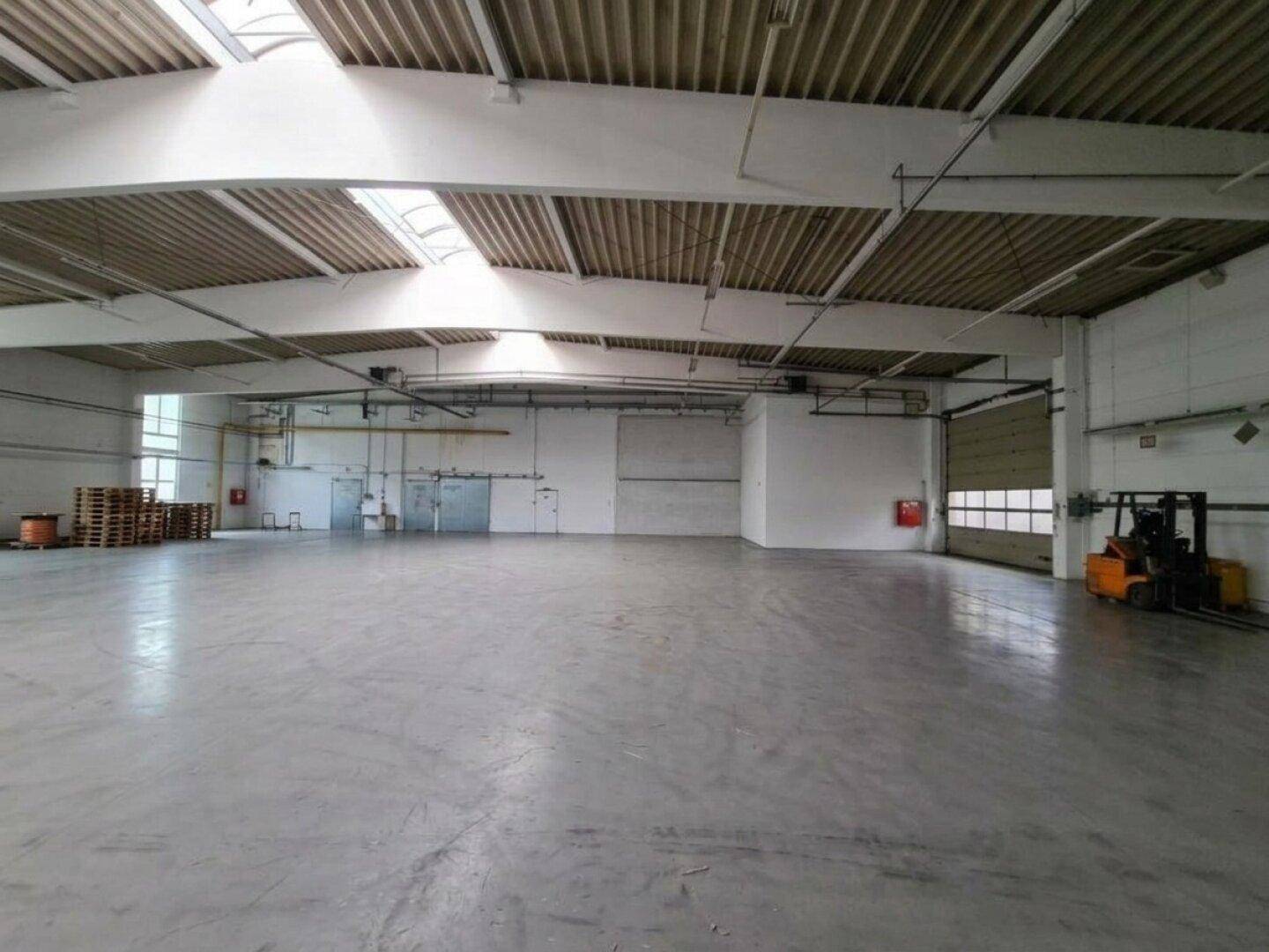Große, leere Lagerhalle mit Betonboden, Rolltoren und einem Gabelstapler am Rand.