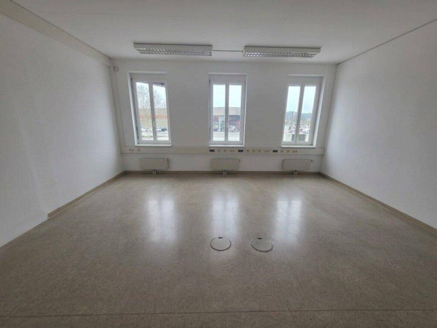 Geräumiger, unmöblierter Büroraum mit drei Fenstern, weißen Wänden und integrierten Bodensteckdosen.
