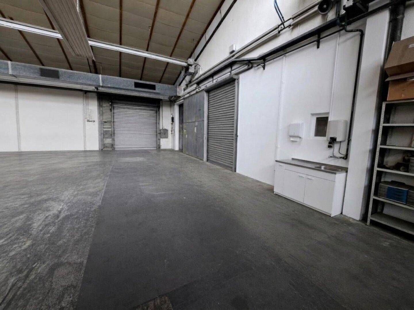 Geräumige Lagerhalle mit hohen Decken, Betonboden und Rolltoren, ausgestattet mit einem Waschbeckenbereich.