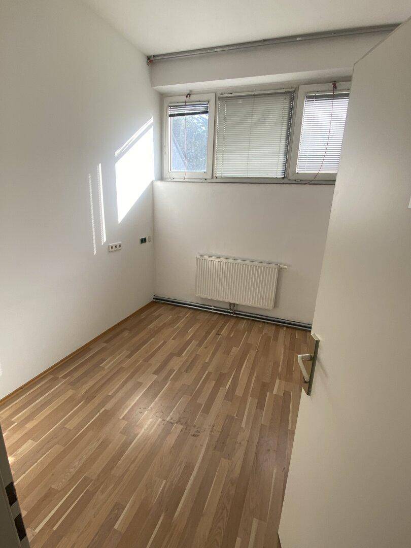 Kleiner, heller Raum mit Holzparkettboden, zwei Fenstern mit Jalousien und einem Heizkörper.