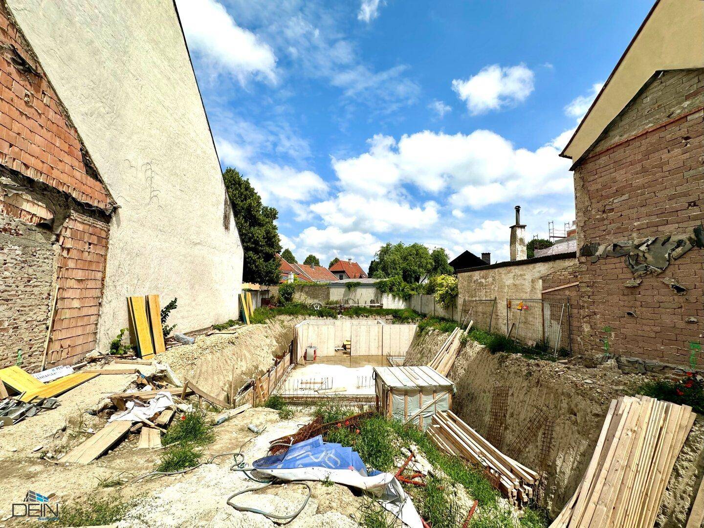 Blick auf die Baugrube mit ersten Fundamentarbeiten und umgebenden Gebäuden an einem sonnigen Tag.