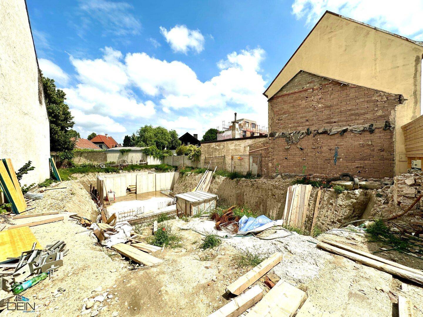 Weitläufige Baustelle mit vorbereiteter Baugrube und angrenzenden Bestandsgebäuden unter blauem Himmel.
