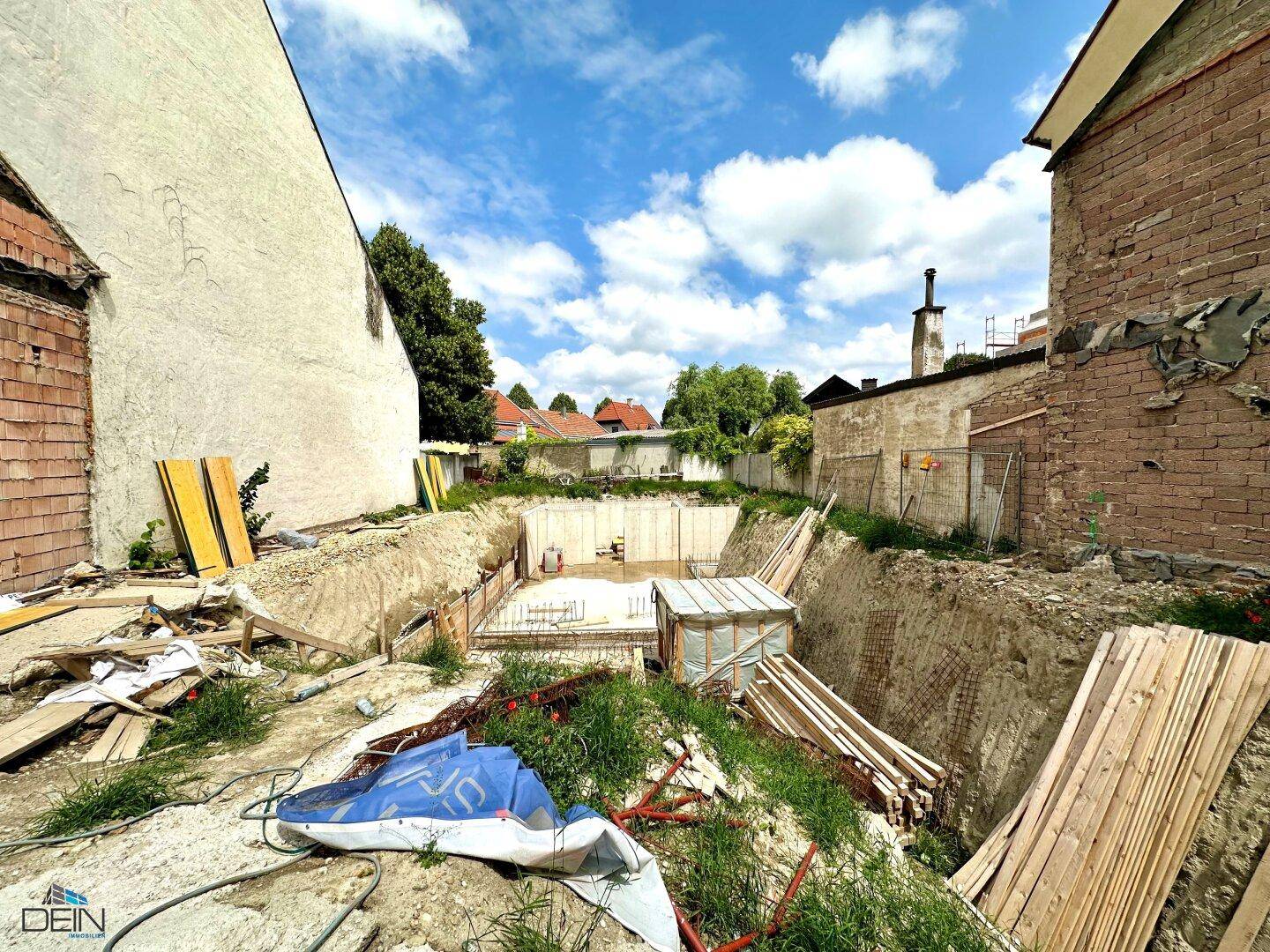 Baustelle mit ausgehobener Grube und vorbereiteten Fundamenten, umgeben von älteren Gebäuden.