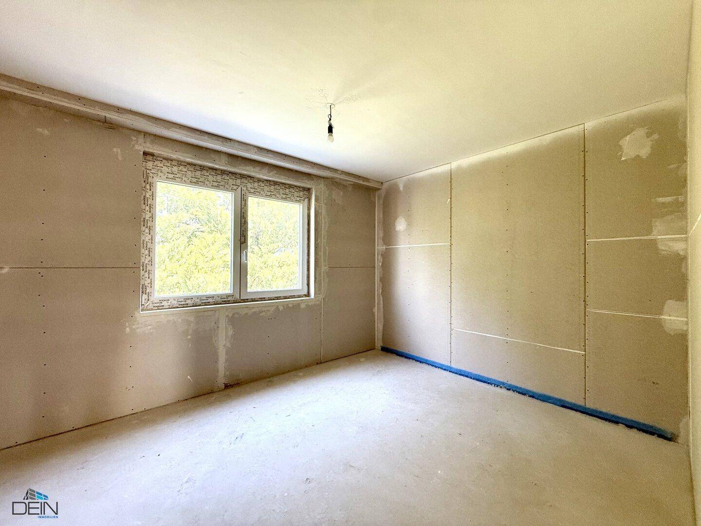 Unfertiger Raum mit Trockenbauwänden und einem großen Fenster, das den Blick ins Grüne freigibt.