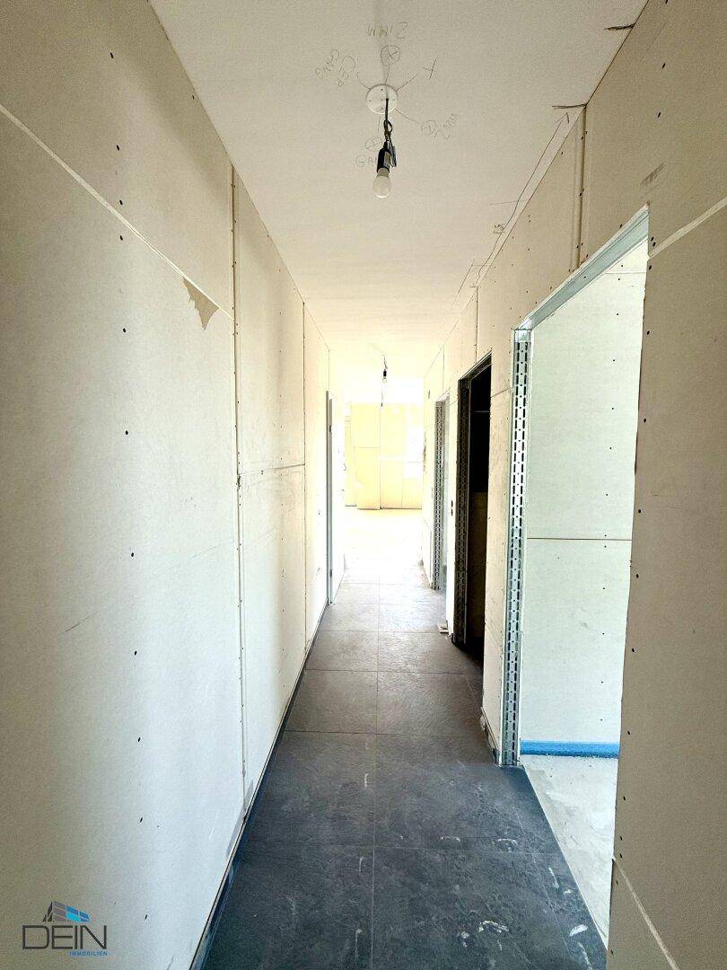 Langer Flur im Rohbauzustand mit Trockenbauwänden und dunklem Boden, der zu weiteren Räumen führt.
