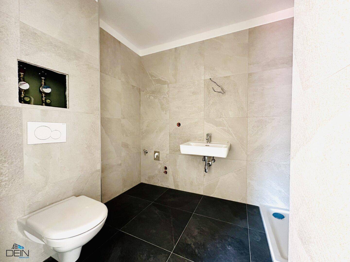 Modernes Badezimmer mit hellgrauen Wandfliesen, dunklem Boden, WC, Waschbecken und Duschbereich.