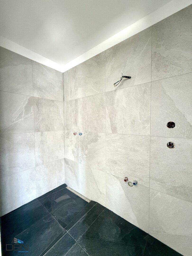 Badezimmer im Bau mit hellgrauen Wandfliesen und dunklem Fliesenboden für die Dusche.