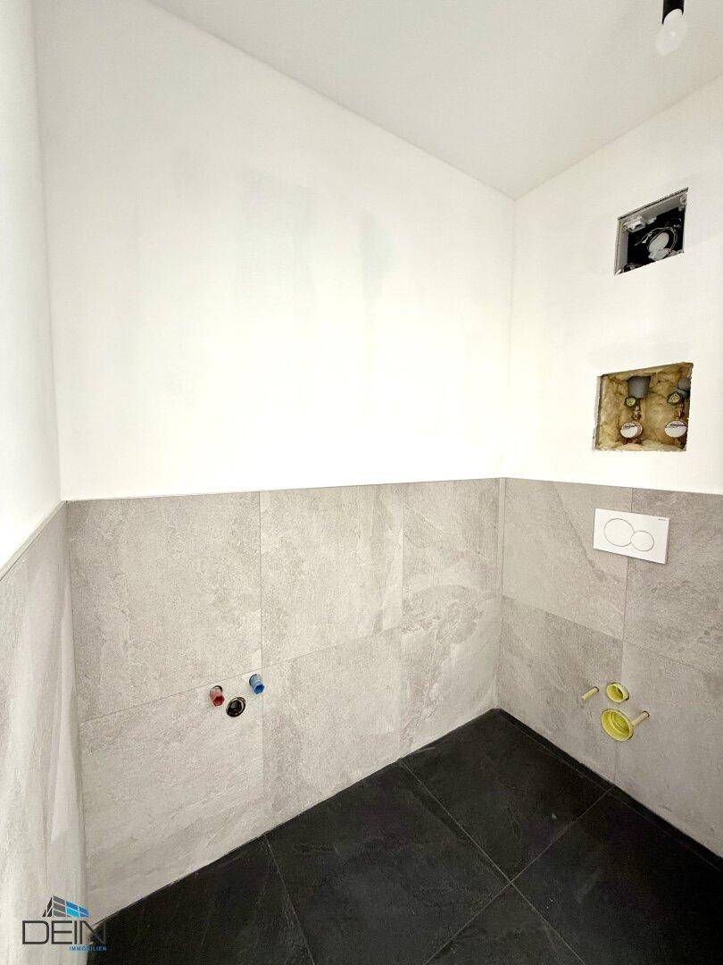 Badezimmer im Rohbau mit grauen Fliesen, vorbereiteten Anschlüssen für WC und Waschbecken.
