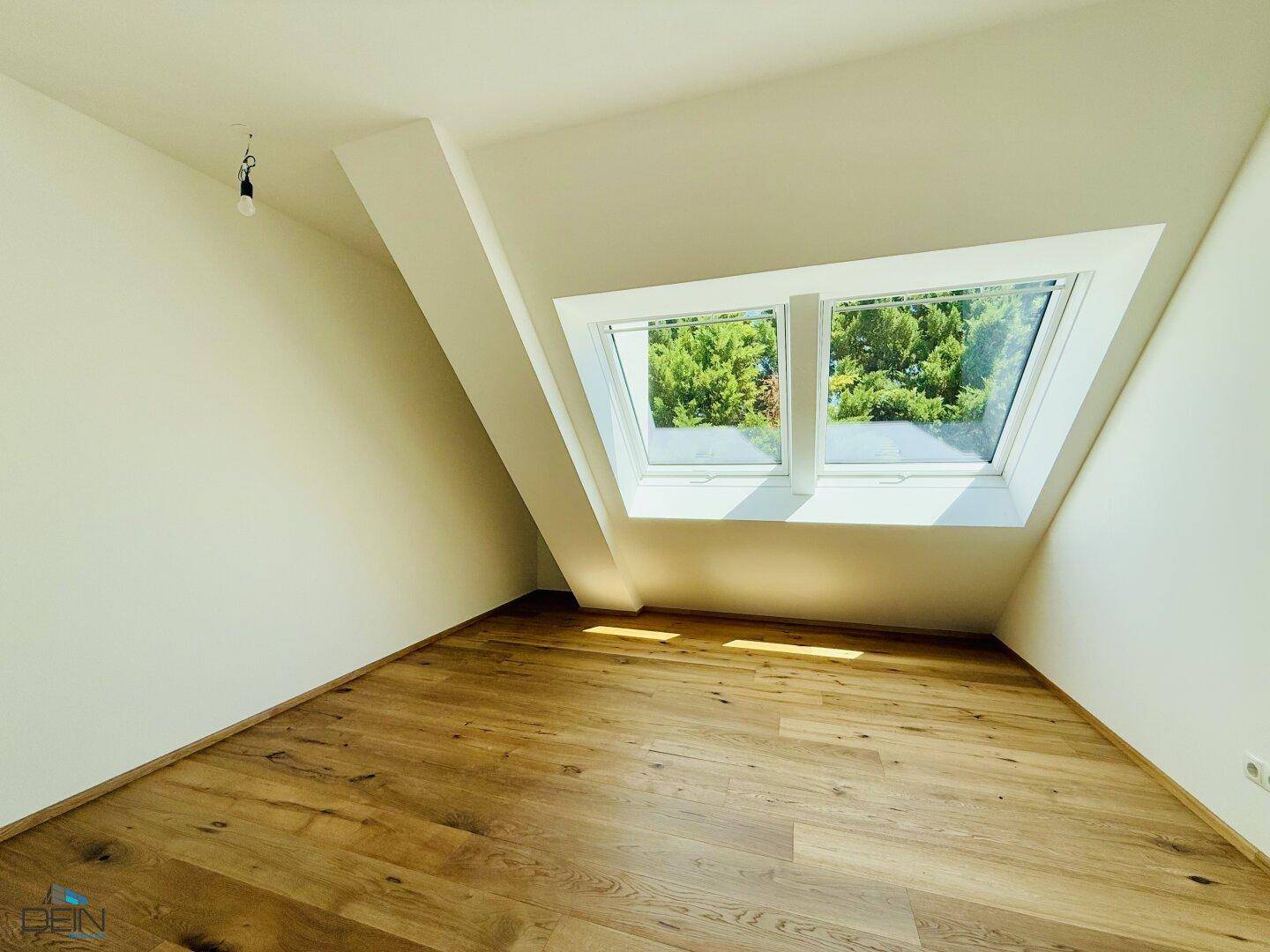 Helles Dachgeschosszimmer mit Holzboden und zwei großen Dachfenstern mit Grünblick.