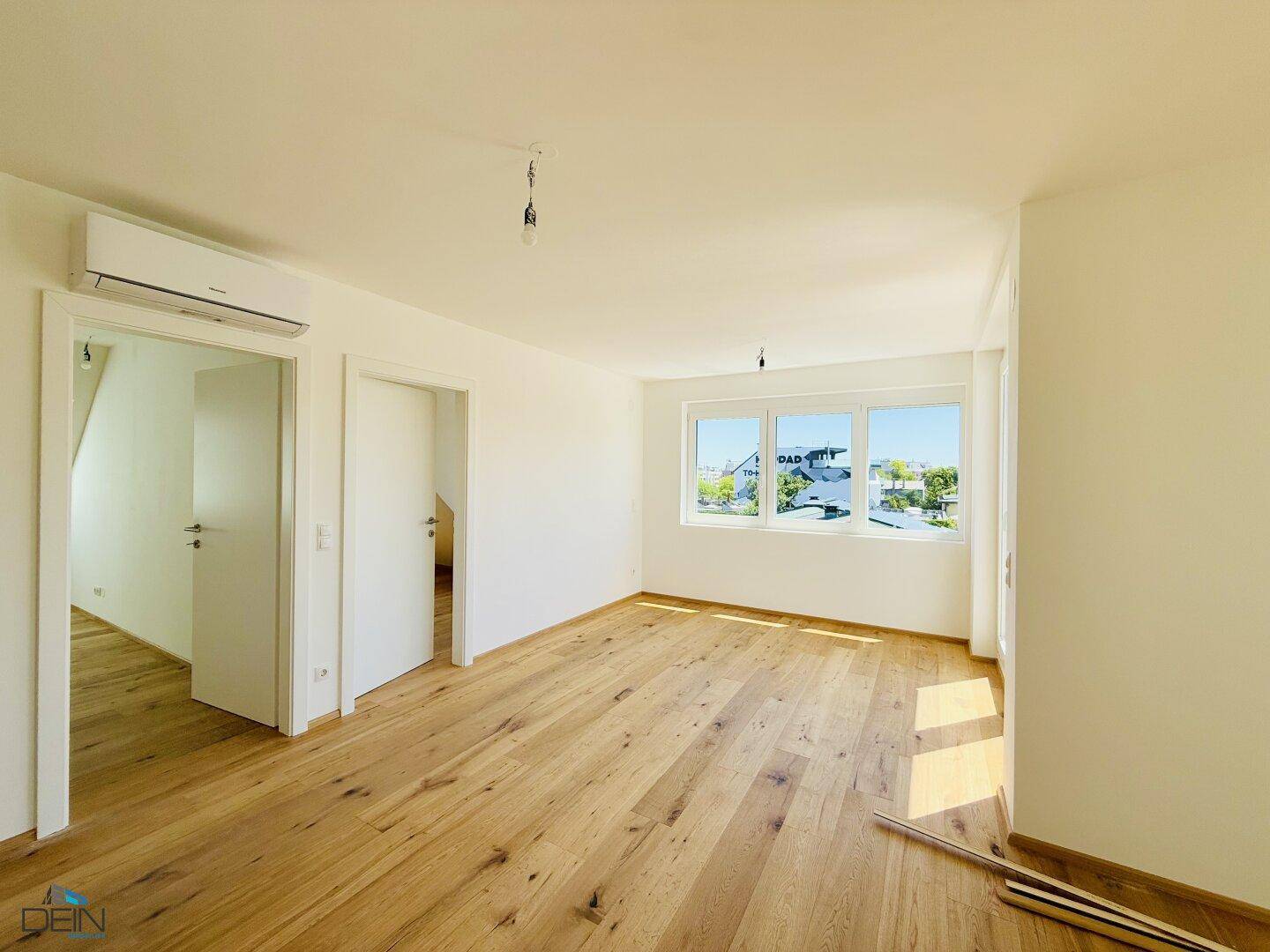 Heller Raum mit Holzboden, Klimaanlage und Blick auf die umliegenden Häuser.