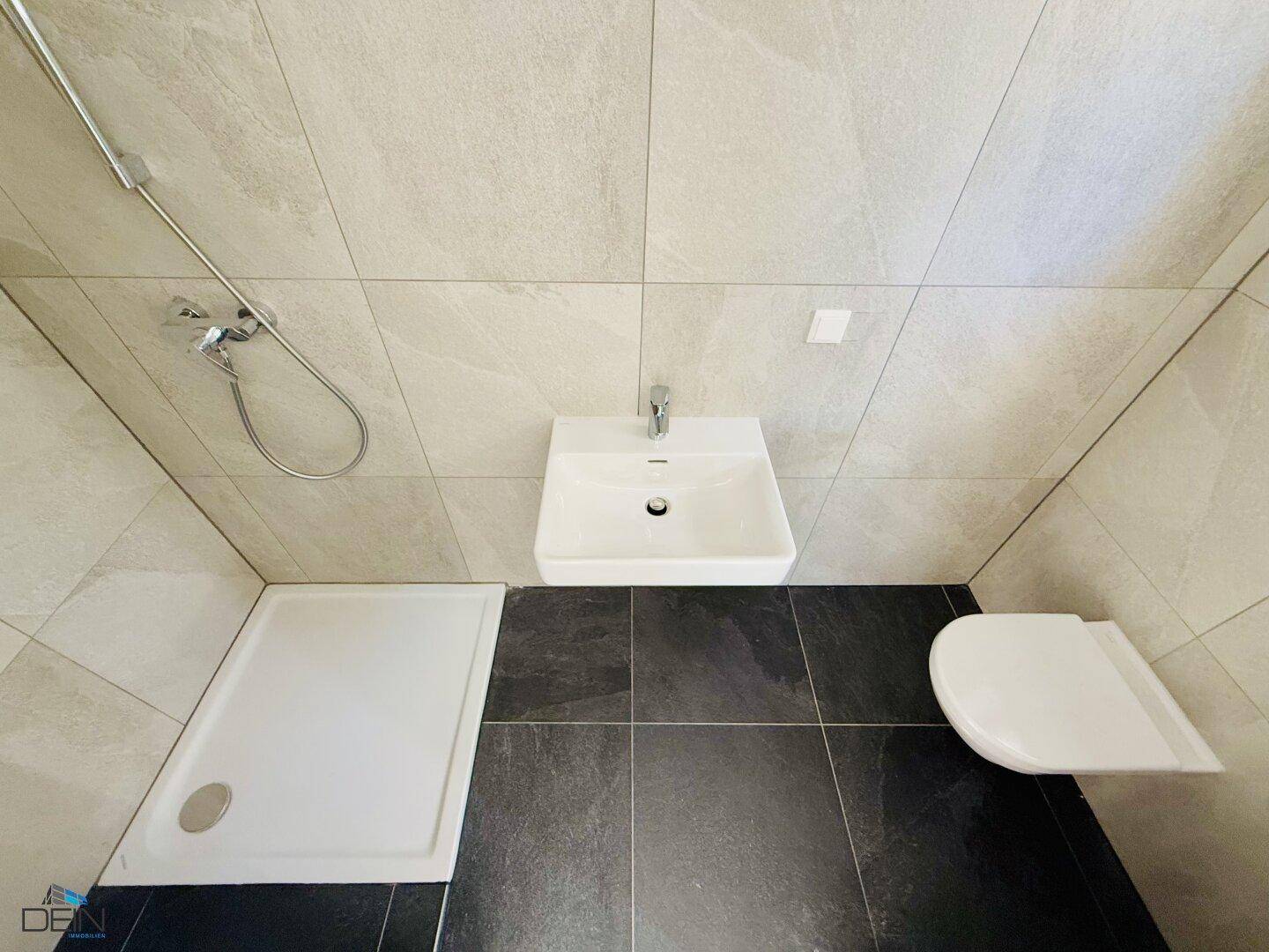 Modernes Badezimmer mit grauen Fliesen, Duschbereich, Wand-WC und kleinem Waschbecken.
