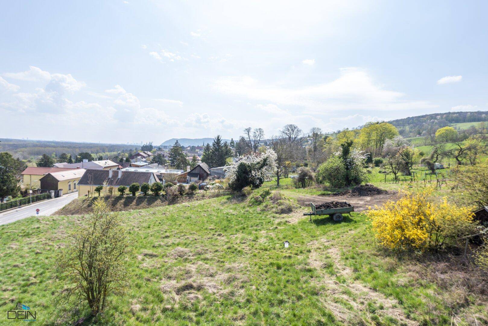 Weitläufiger Blick auf ein Dorf in der Ferne, umgeben von grünen Wiesen und Bäumen.