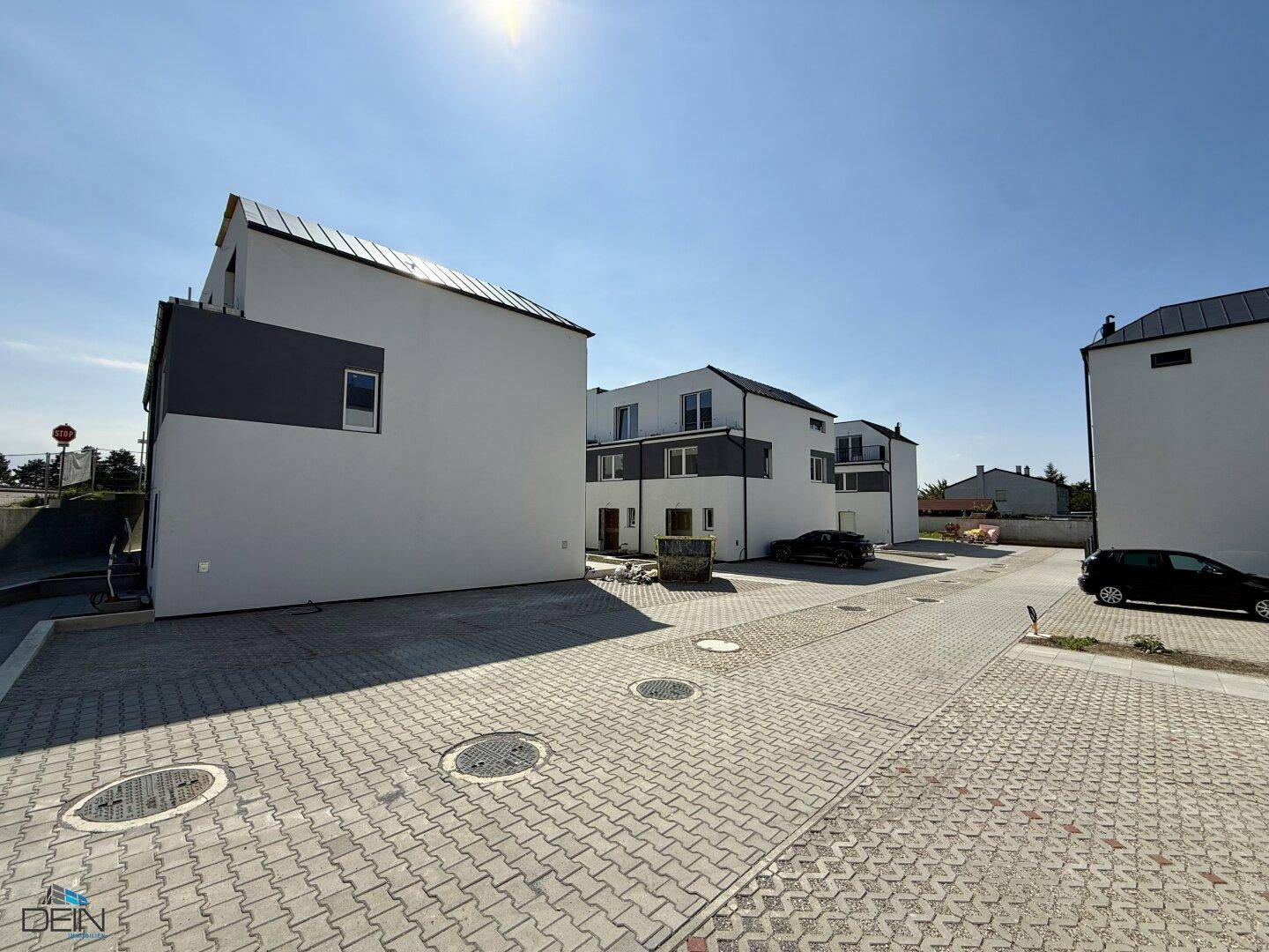 Seitenansicht eines modernen Reihenhauses mit weißer Fassade und gepflastertem Vorplatz an einem sonnigen Tag.