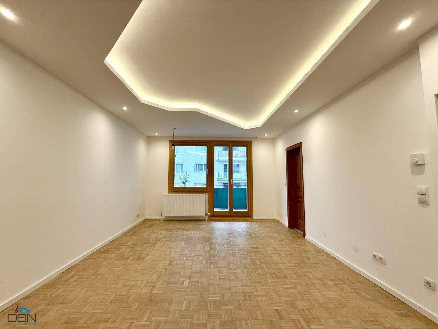 Geräumiges Wohnzimmer mit Parkettboden, moderner Deckenbeleuchtung und Blick nach draußen.