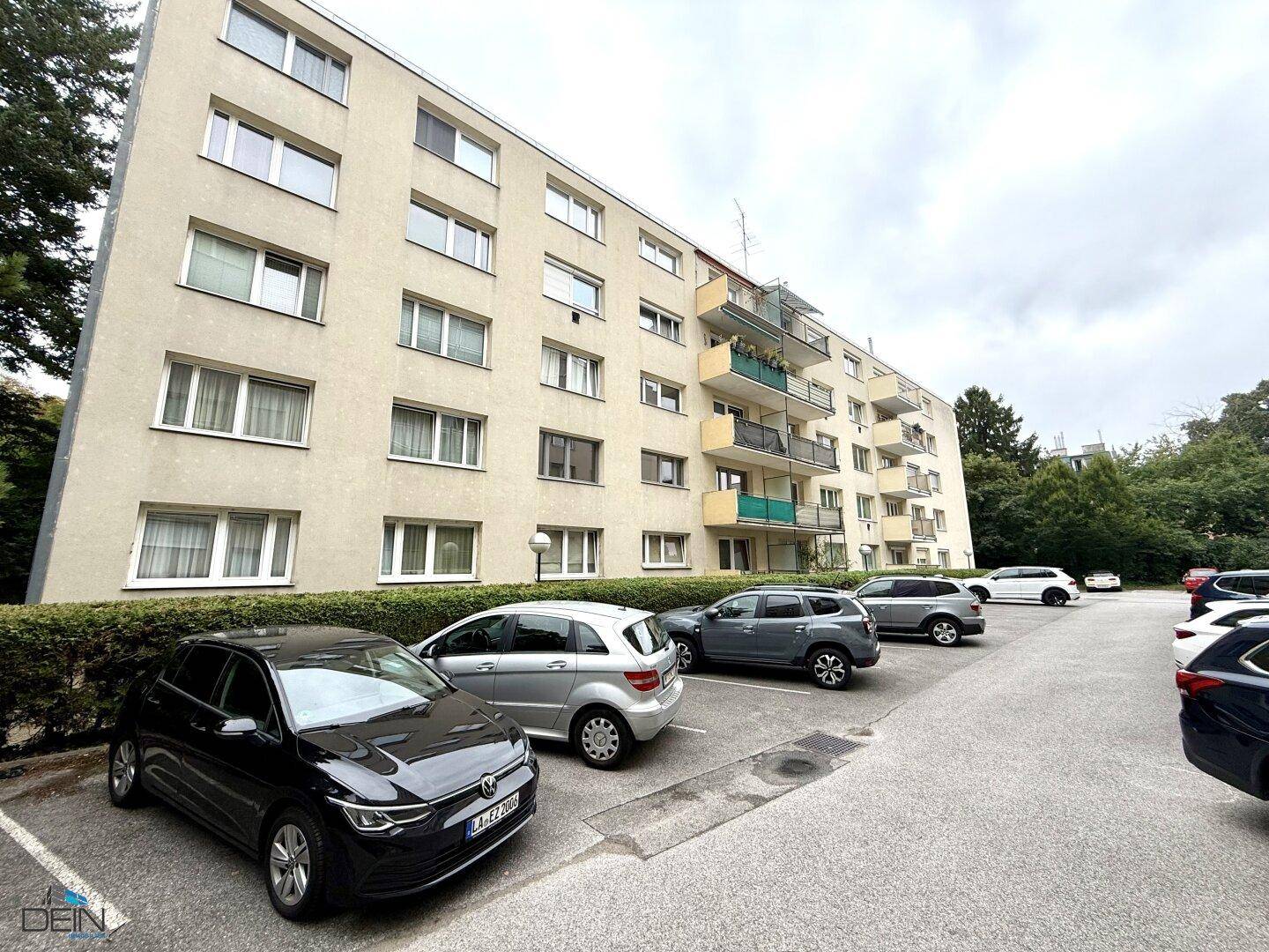 Mehrfamilienhaus mit Parkplätzen im Freien und grüner Umgebung.