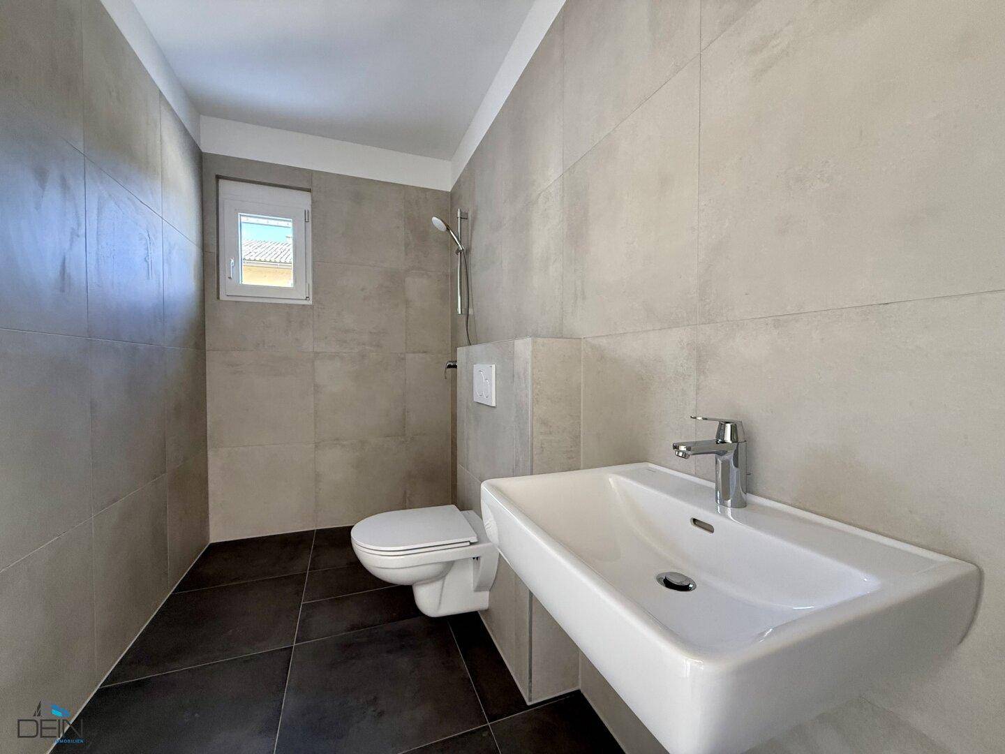 Modernes Badezimmer mit Wand-WC, Waschbecken und Duschbereich, hell gefliest und mit Fenster.