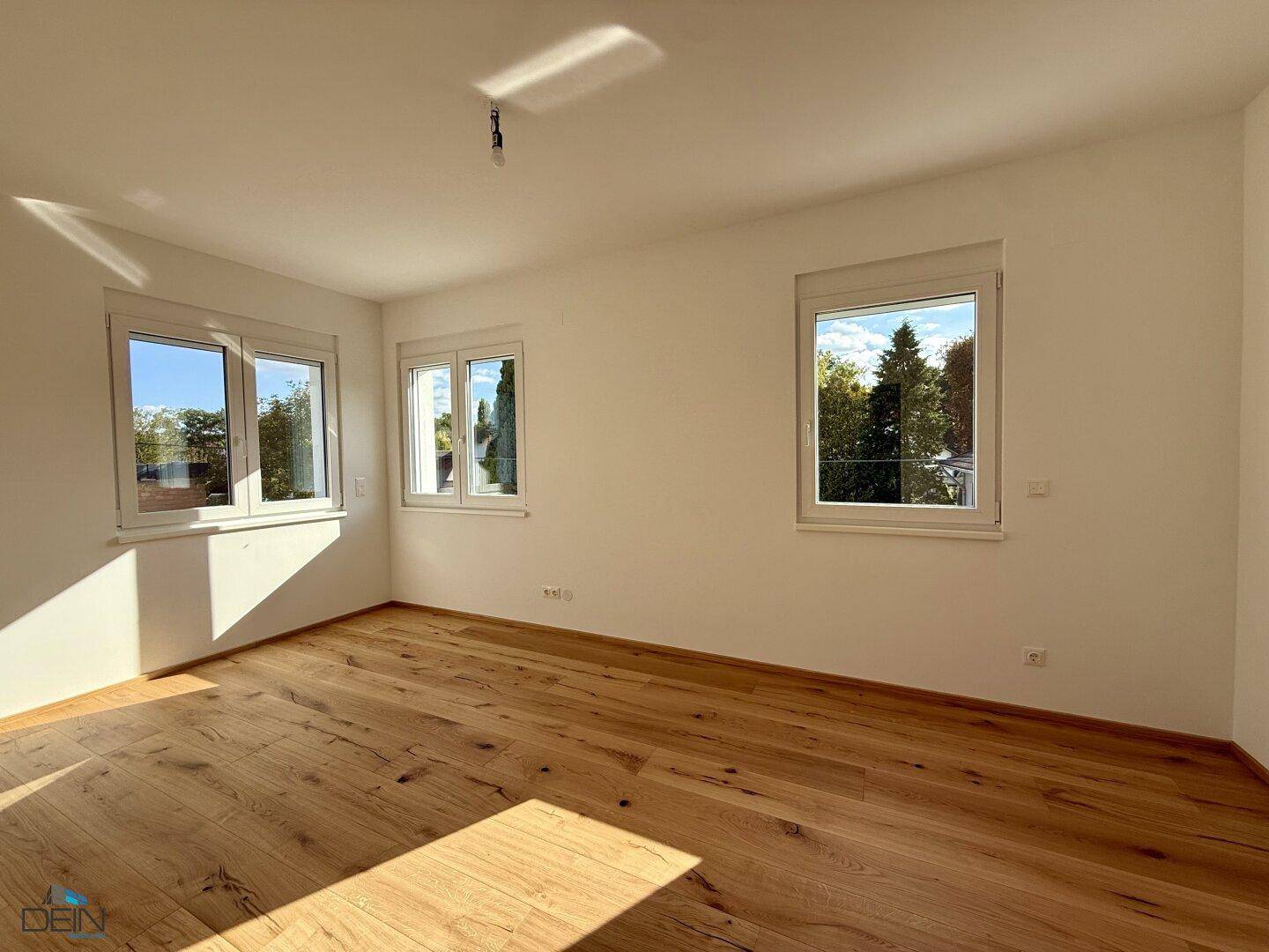 Helles Schlafzimmer mit Holzboden und mehreren Fenstern, die viel Sonnenlicht hereinlassen und eine grüne Aussicht bieten.