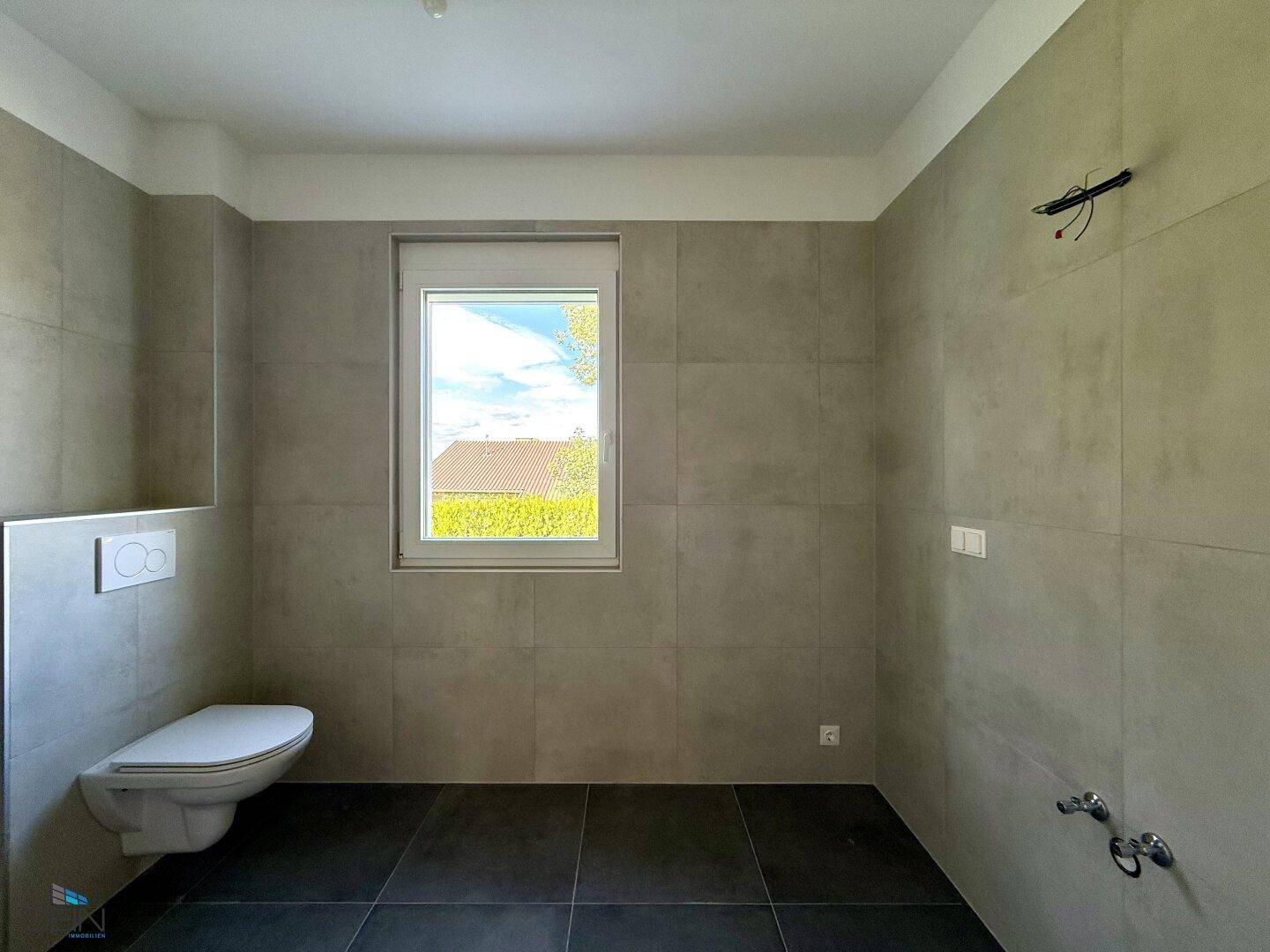 Modernes WC mit Wand-WC und Fenster, in hellen Grautönen gefliest und funktional eingerichtet.