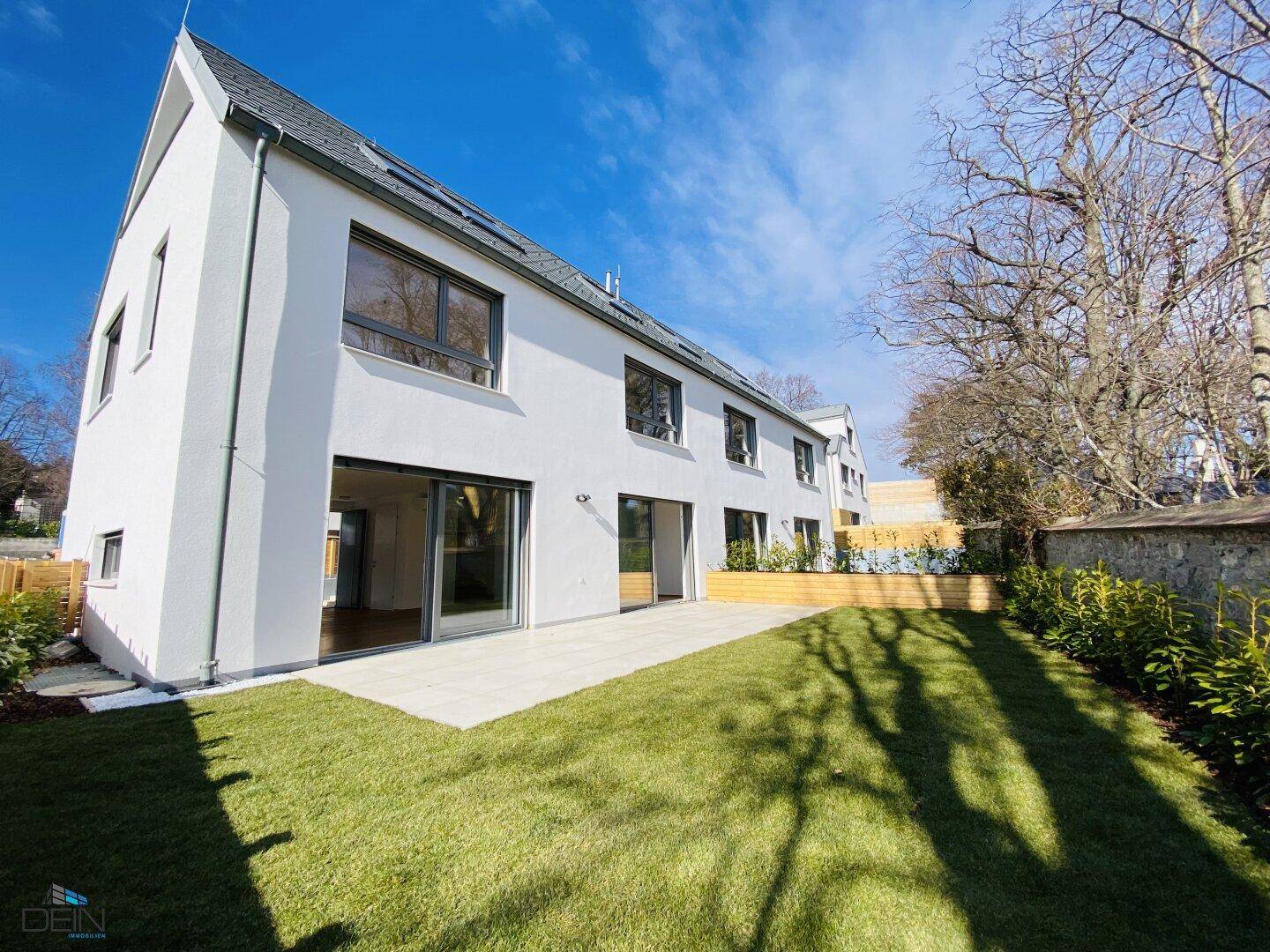 Moderne weiße Hausfassade mit großer Terrasse und gepflegtem Rasen unter blauem Himmel.