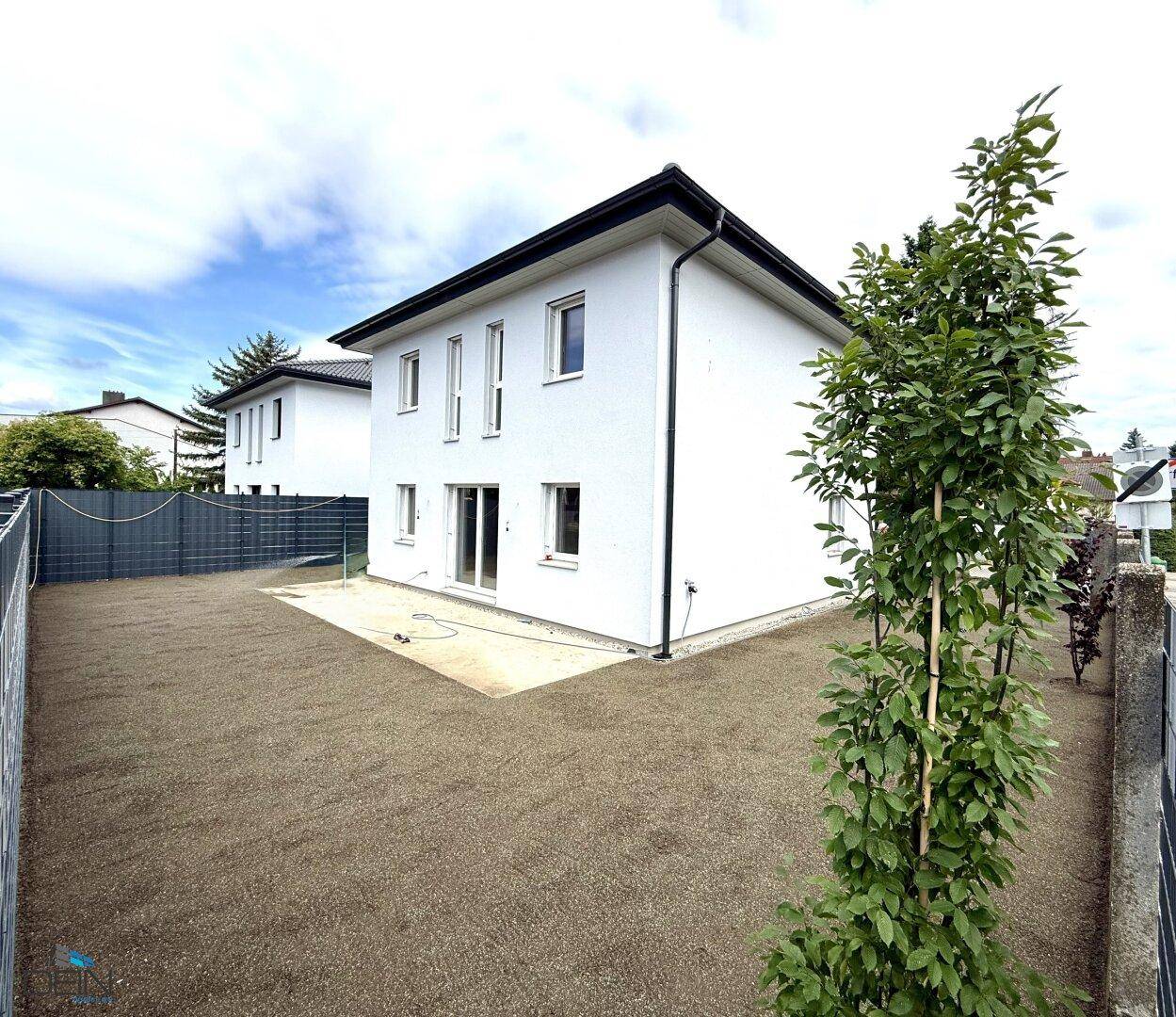 Neubau-Einfamilienhaus mit vorbereitetem Gartenbereich und kleiner Terrasse unter bewölktem Himmel.