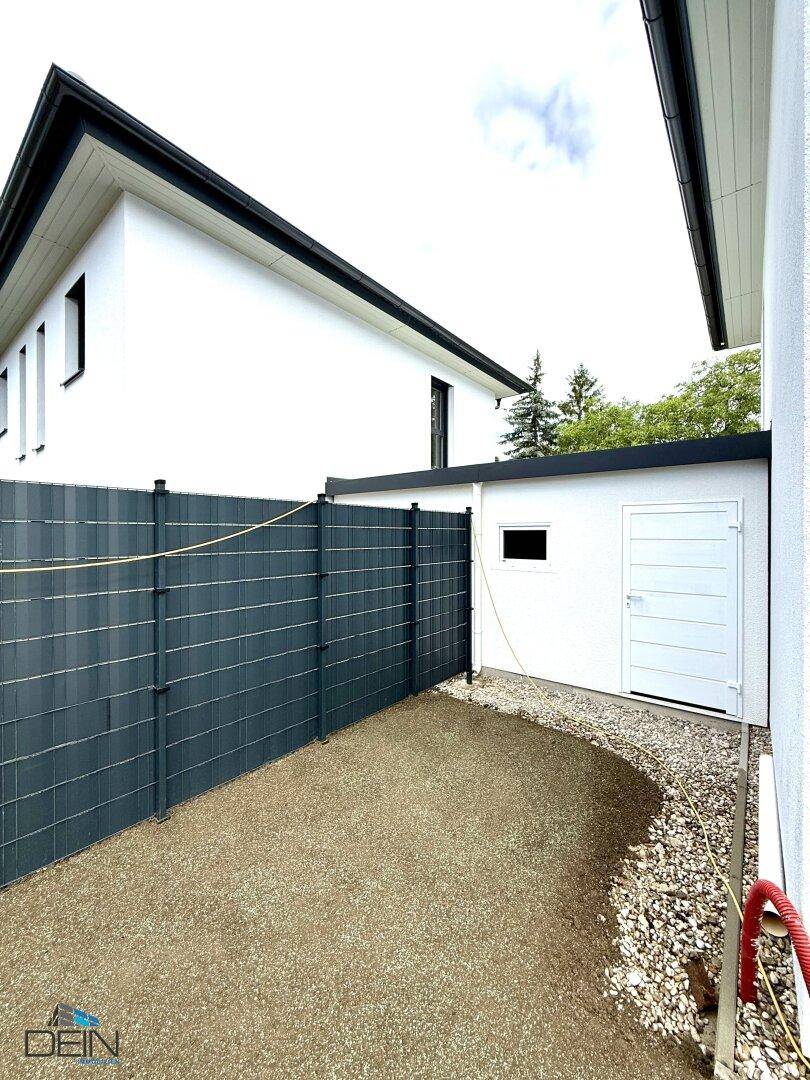 Seitenansicht des Hauses mit einem kleinen Nebengebäude und einem Zaun, der das Grundstück begrenzt.
