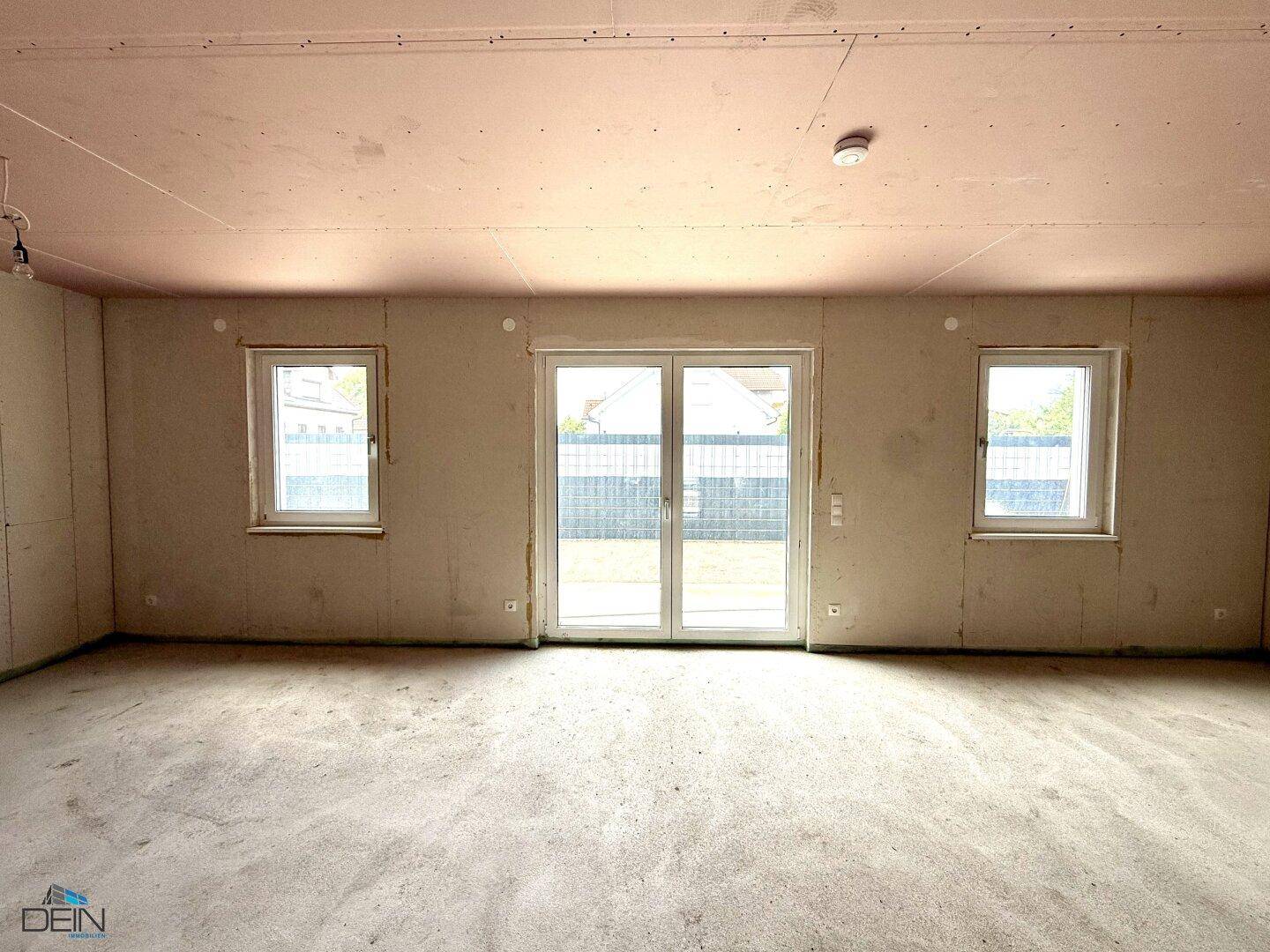 Rohbau-Wohnbereich mit großen Fenstern und Terrassentür, die viel Tageslicht hereinlassen.