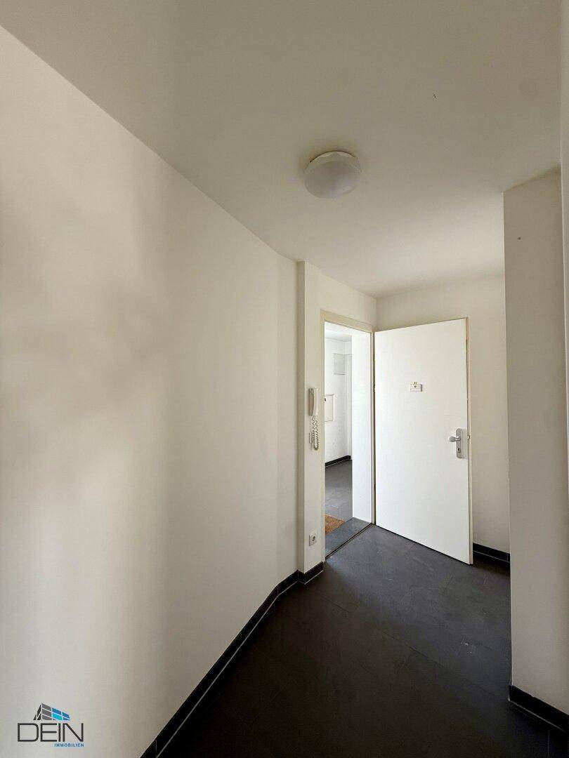Heller Flur mit dunklem Fliesenboden und weißen Wänden, der den Blick in einen angrenzenden Raum freigibt.