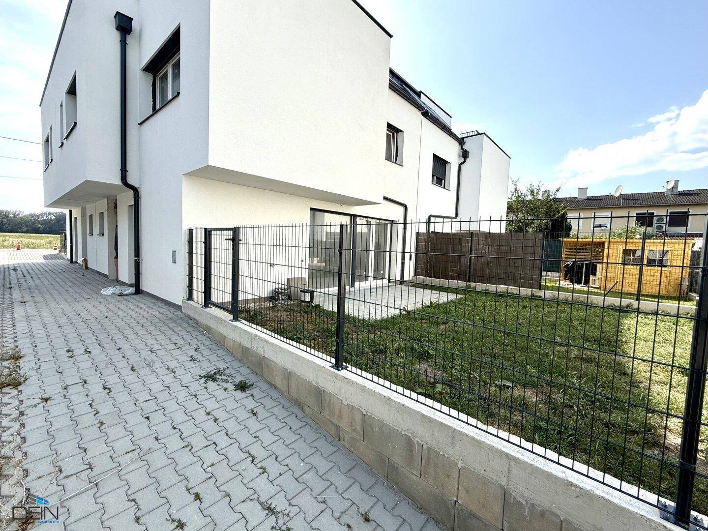 Gartenbereich mit Rasenfläche und gepflasterter Terrasse, umgeben von einem Zaun.