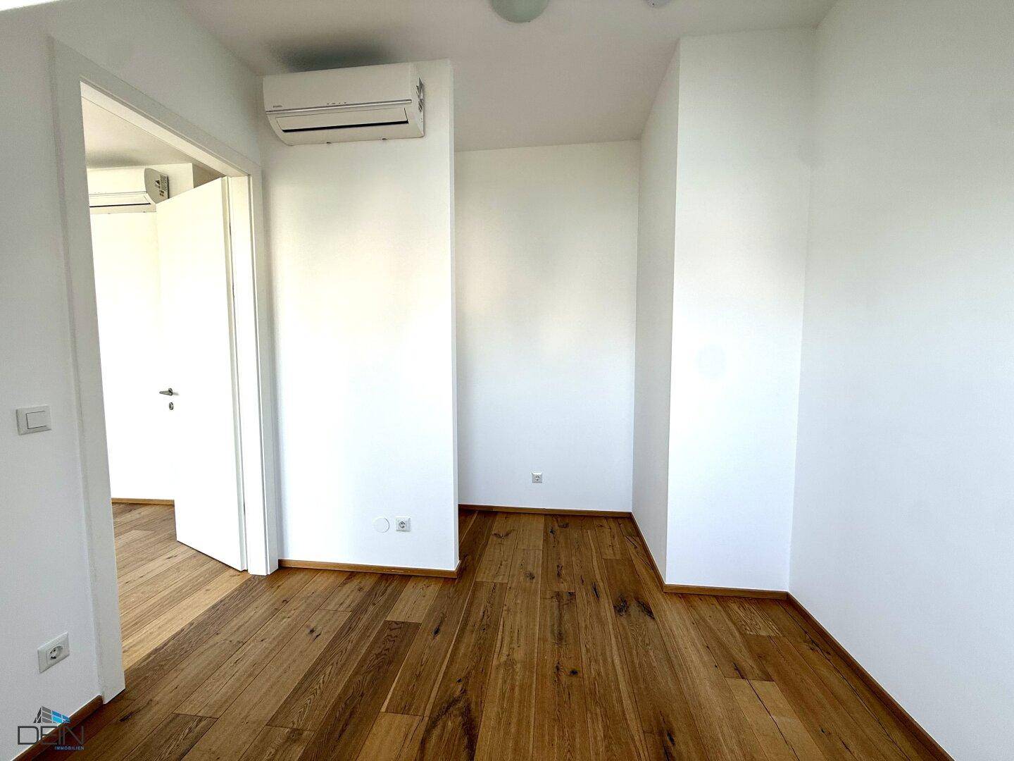 Heller Wohnbereich mit Holzboden und Klimaanlage, der Zugang zu einem weiteren Zimmer bietet.