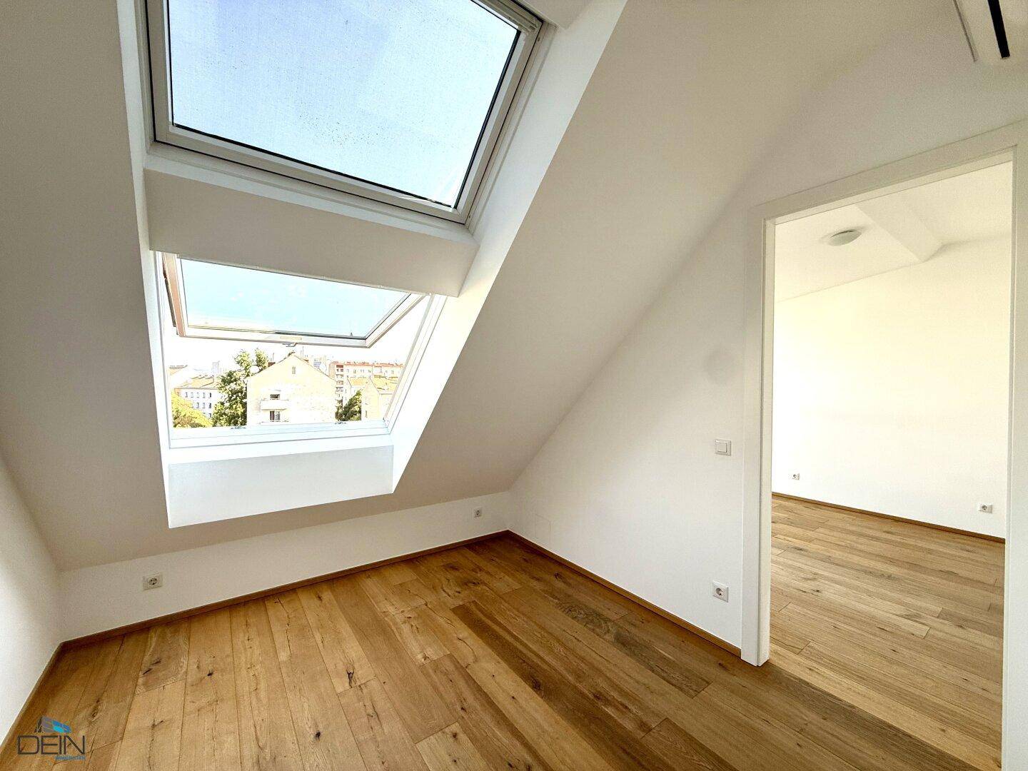 Helles Zimmer mit Holzboden und einem großen Dachfenster, das einen Blick nach draußen bietet.