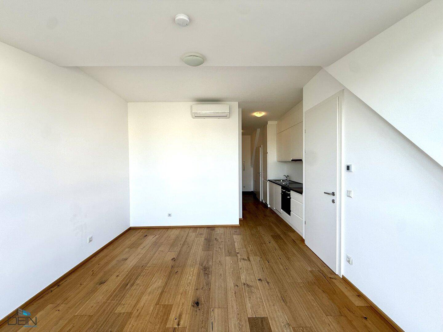 Heller Wohnbereich mit Holzboden und Klimaanlage, im Hintergrund eine moderne Küchenzeile.