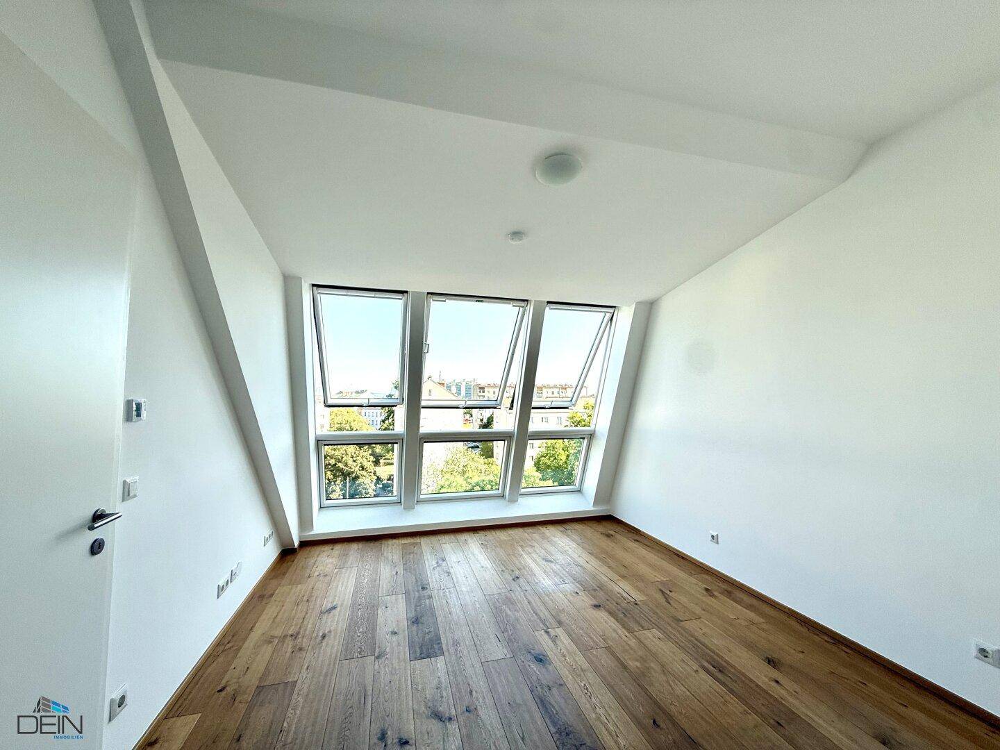 Geräumiges Zimmer im Dachgeschoss mit Holzboden und schrägen Wänden, durchflutet von Tageslicht.