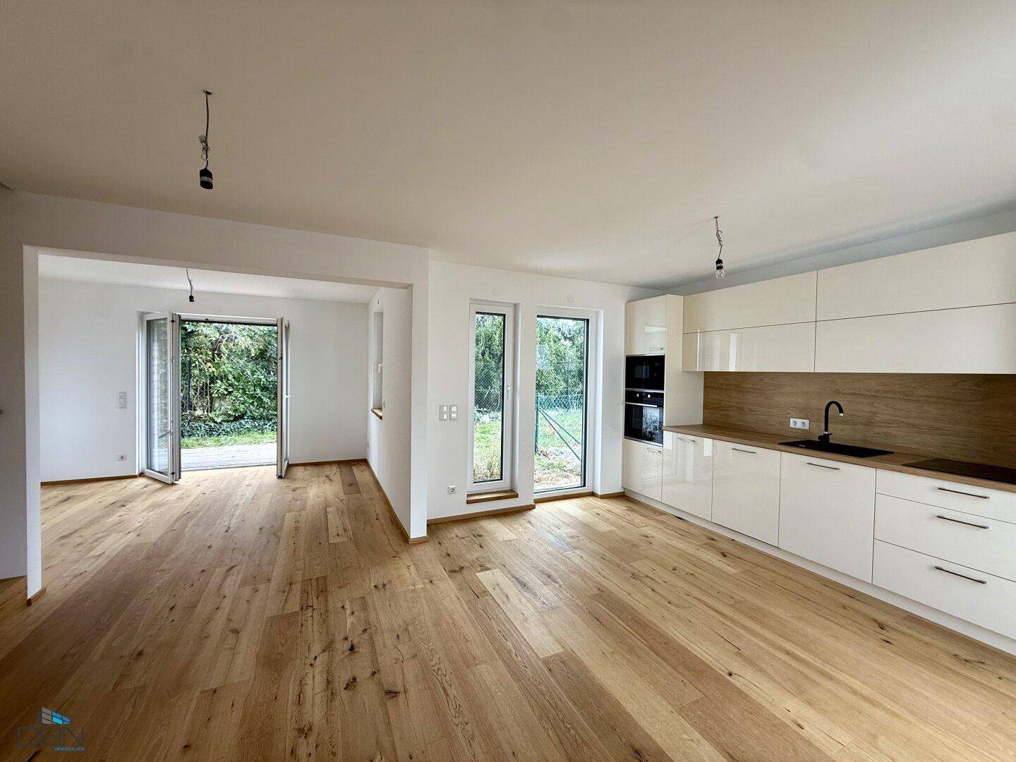 Geräumiger Wohnbereich mit integrierter Küche, Holzboden und großen Fenstern zum Garten.