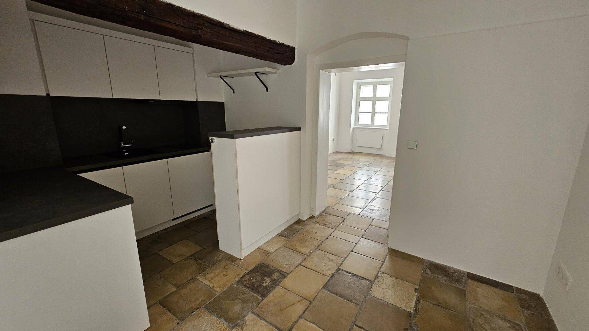 Kleine Küche mit weißen Schränken, dunklen Arbeitsplatten und freiliegendem Holzbalken, offen zum nächsten Raum.