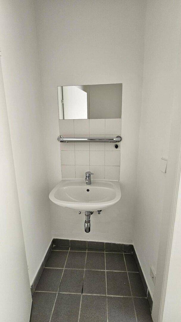 Waschbecken mit Spiegel und Handtuchhalter, umgeben von weißen Fliesen und dunklem Boden.