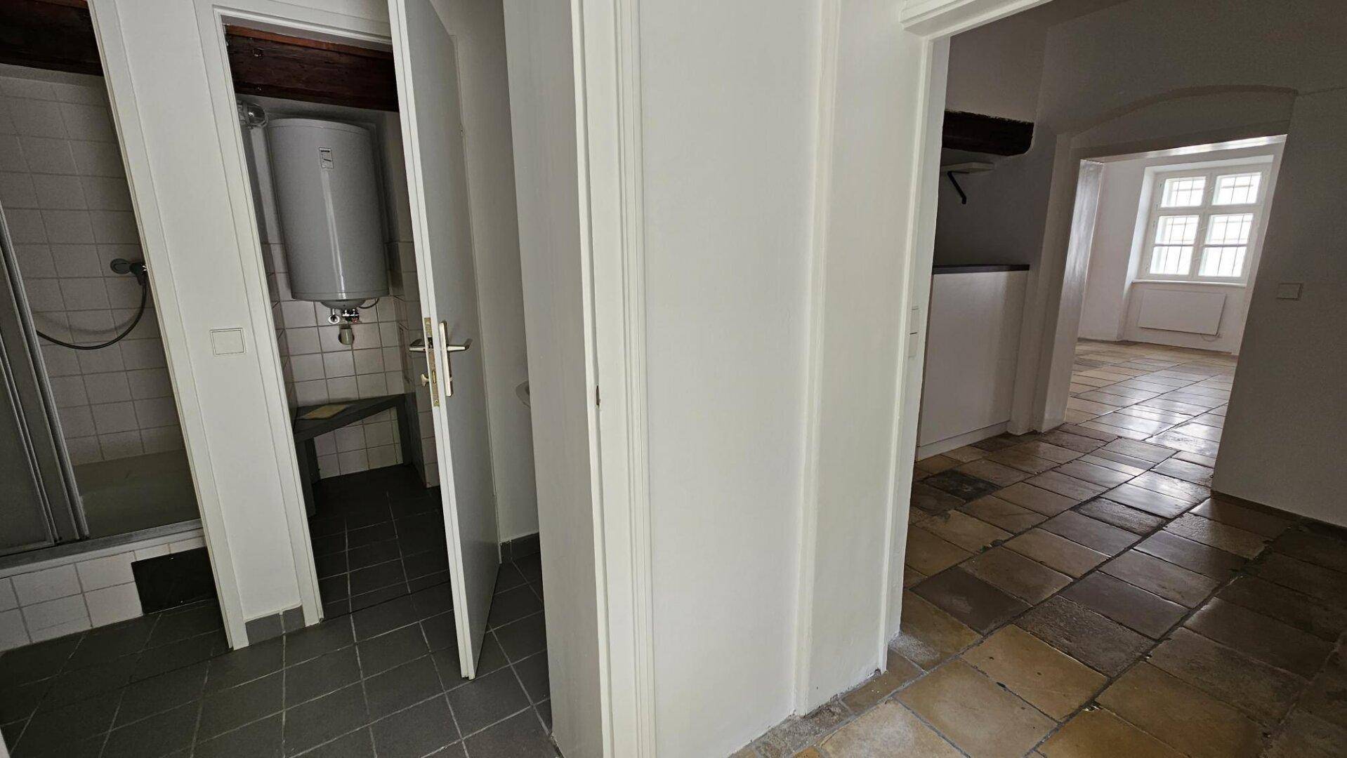 Eingangsbereich zu einem kleinen Badezimmer mit Duschkabine und Warmwasserboiler, funktional gestaltet.